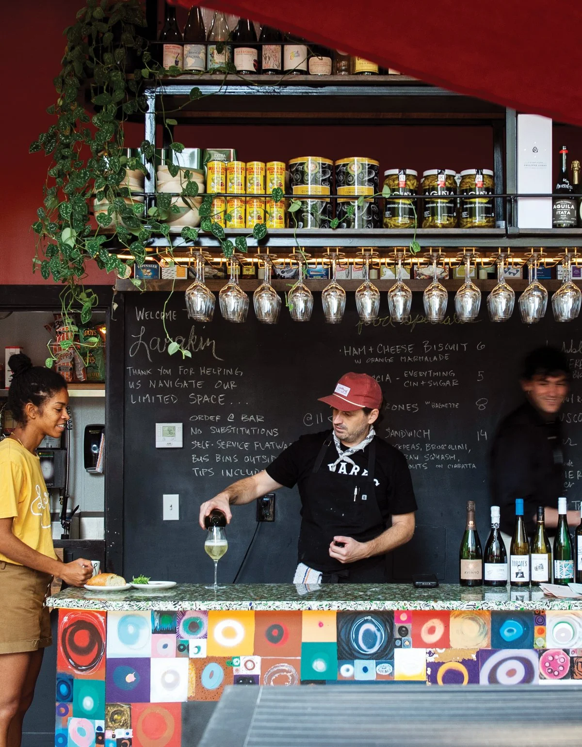A bar scene with a woman on the left ordering at the counter, a bartender pouring wine in the center, and another person smiling in the background. The bar has colorful abstract artwork on the front, hanging wine glasses, and shelves with bottles and canned goods. A black chalkboard menu is on the wall behind them.
