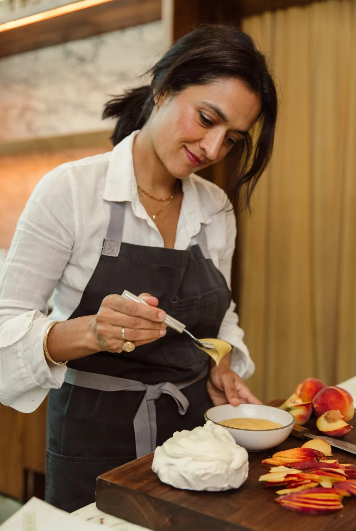 A woman in a white shirt and black apron preparing a dessert with peaches and whipped cream in a cozy kitchen.