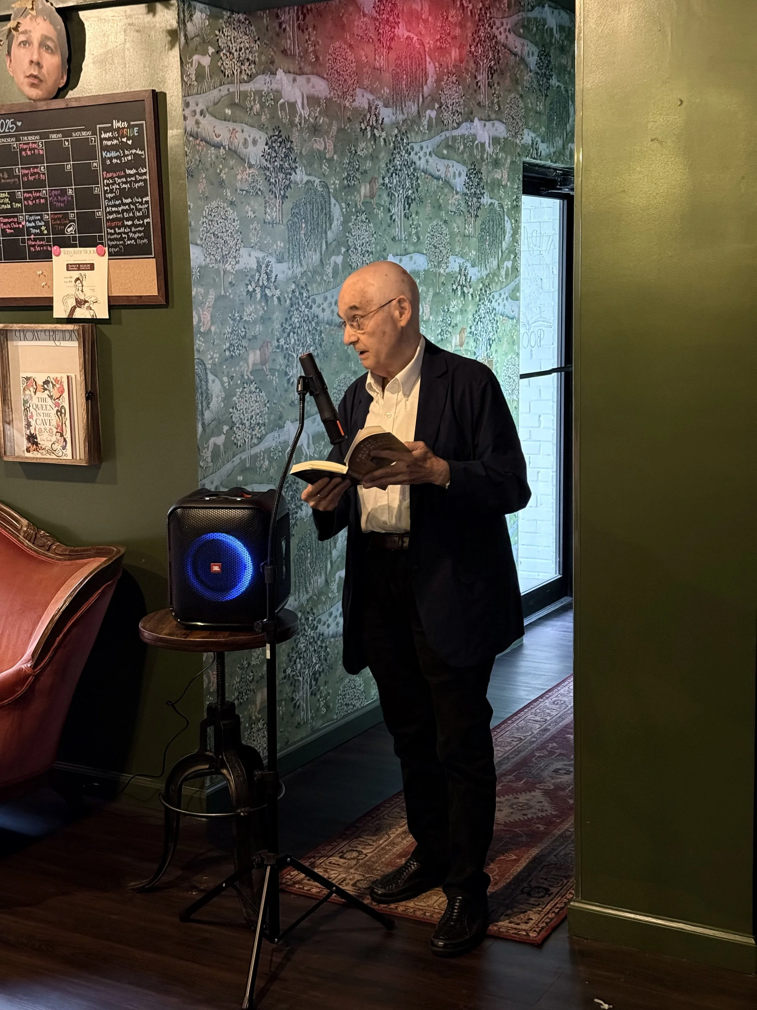 A man with glasses in a dark suit reading a book into a microphone in a room with green walls, a decorative tapestry, a wooden chair, and a black JBL speaker.