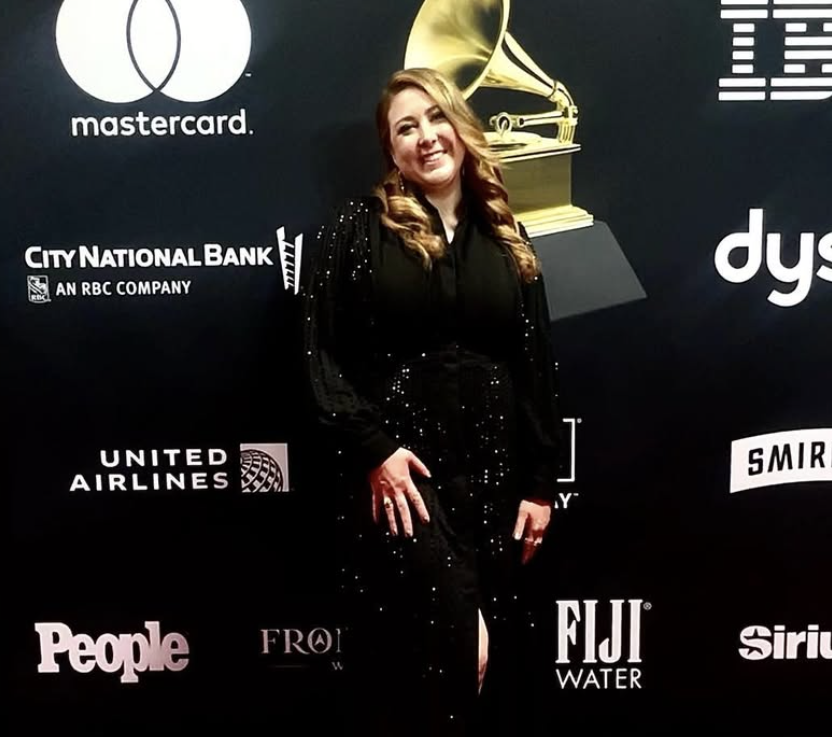 A woman with long, wavy blonde hair wearing a black sequined dress posing in front of a black backdrop with various sponsor logos and a large golden Grammy statue.