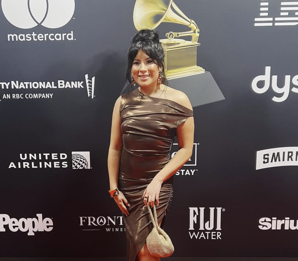 Woman in a shiny brown dress holding a small, beaded handbag on a red carpet at an event with logos of Mastercard, United Airlines, People, and other sponsors on the backdrop.