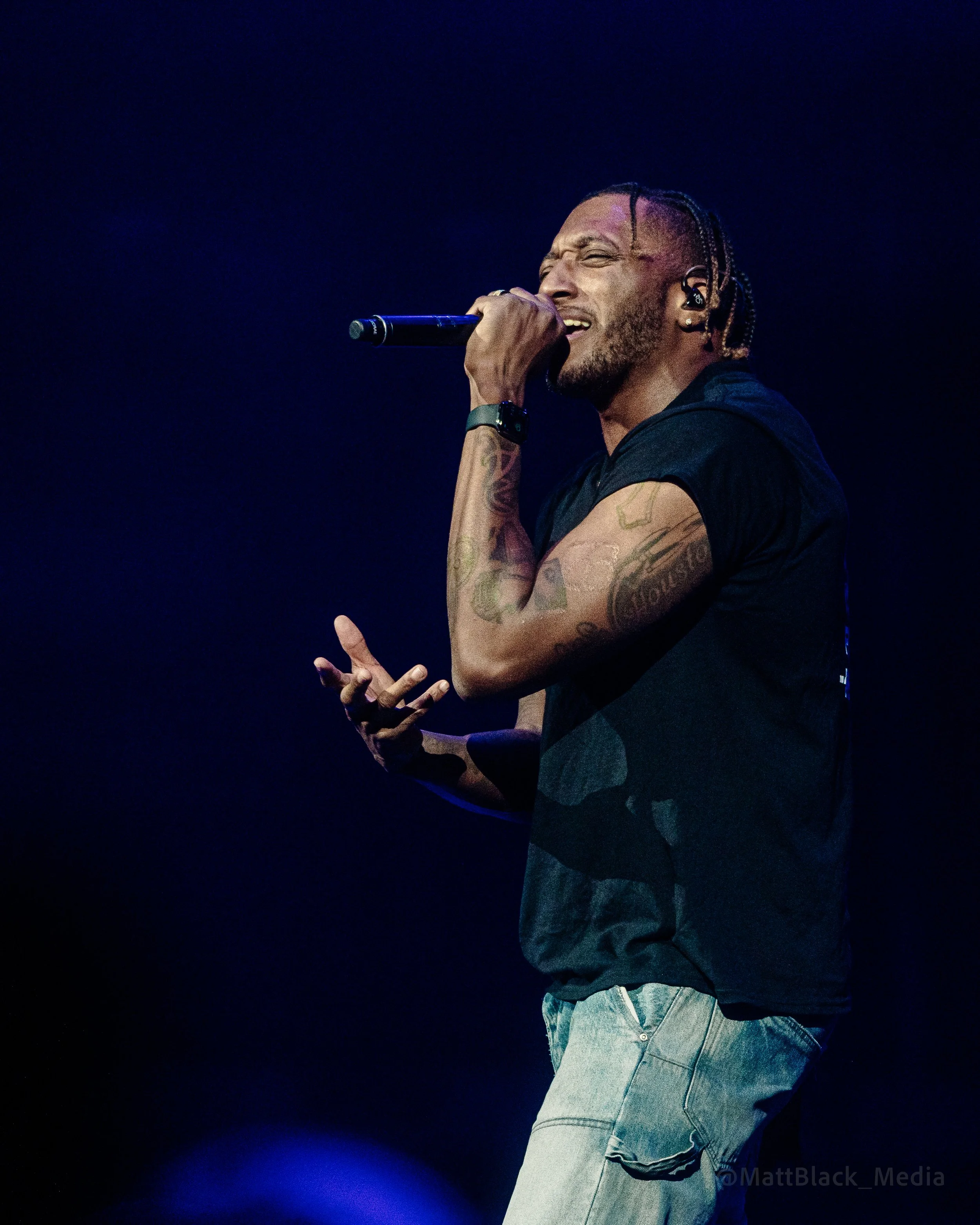 A male singer performing on stage, holding a microphone close to his mouth, with a dark background and stage lighting.