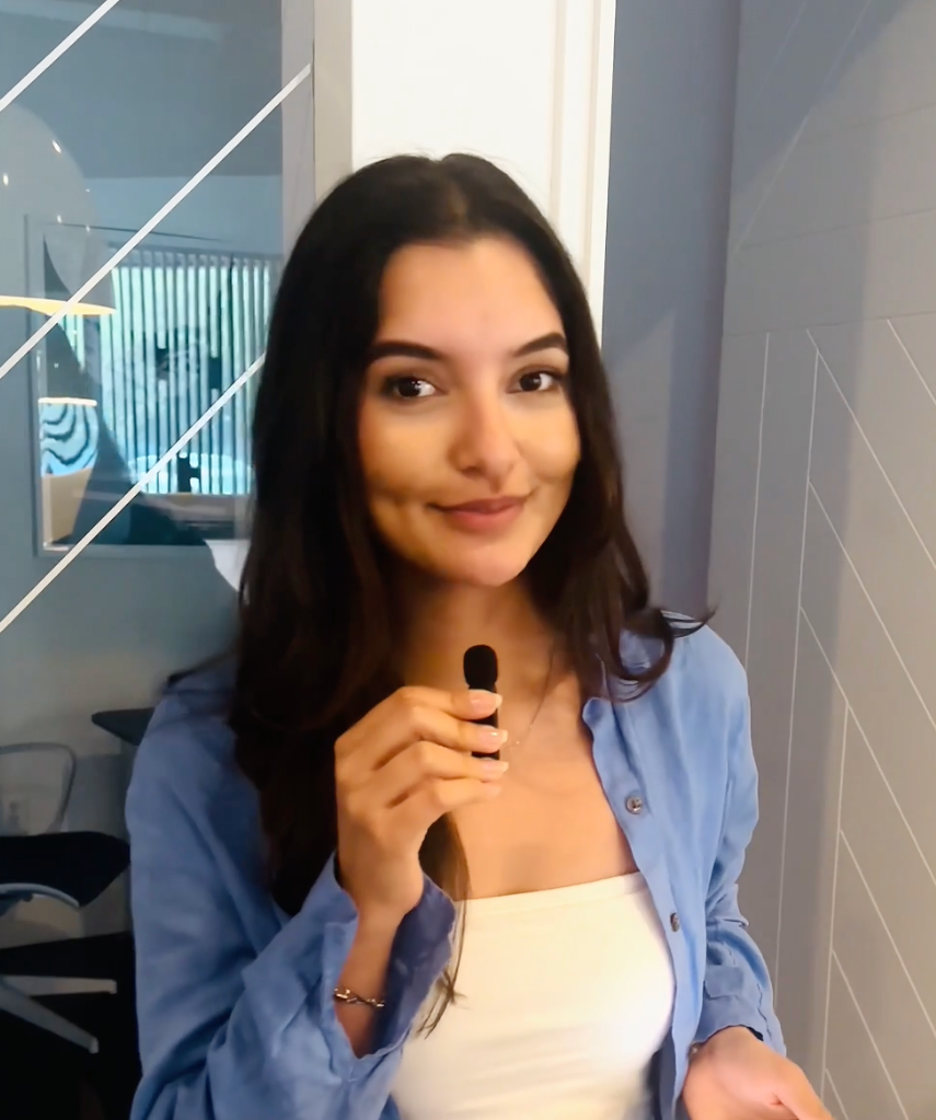 A young woman with long dark hair holding a microphone in an indoor setting, smiling and wearing a light blue shirt over a white top.