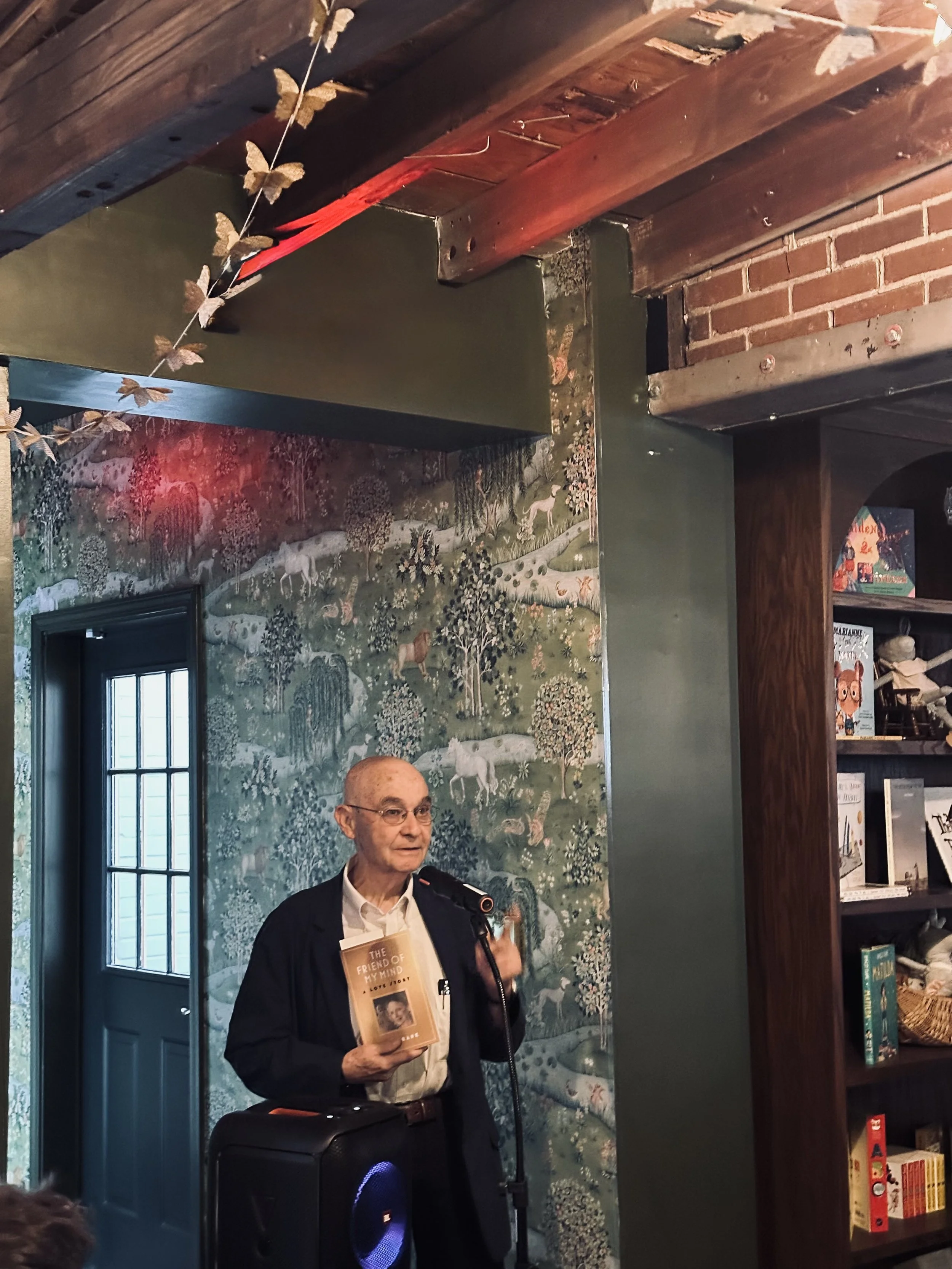 An elderly man with glasses and a bald head holding a book titled 'The Friend of My Mind' and speaking into a microphone, standing in front of a wall with a nature-themed mural and bookshelves.