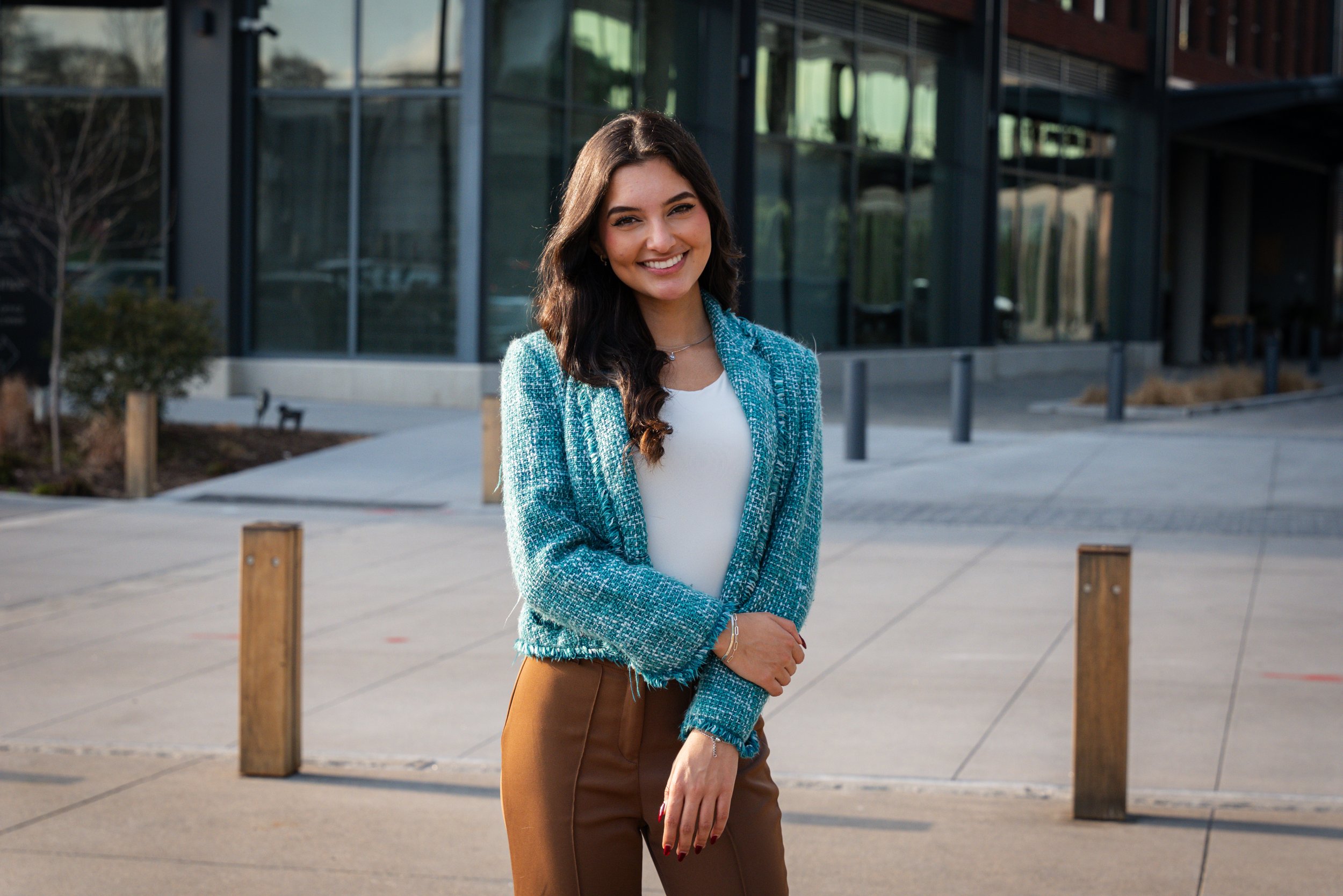 A smiling woman with long dark hair, wearing a teal jacket, white top, and brown pants, standing outdoors in front of a modern glass building during daytime.