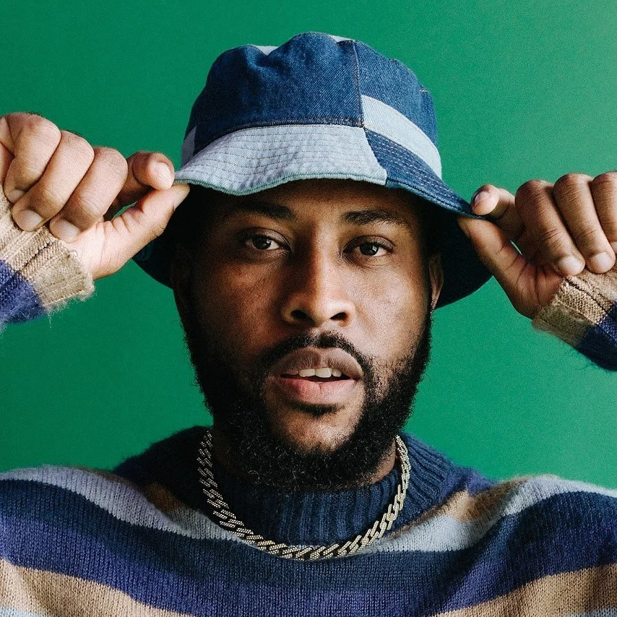 Close-up portrait of a man wearing a patchwork bucket hat and a striped sweater, holding the brim of his hat with both hands, against a green background.
