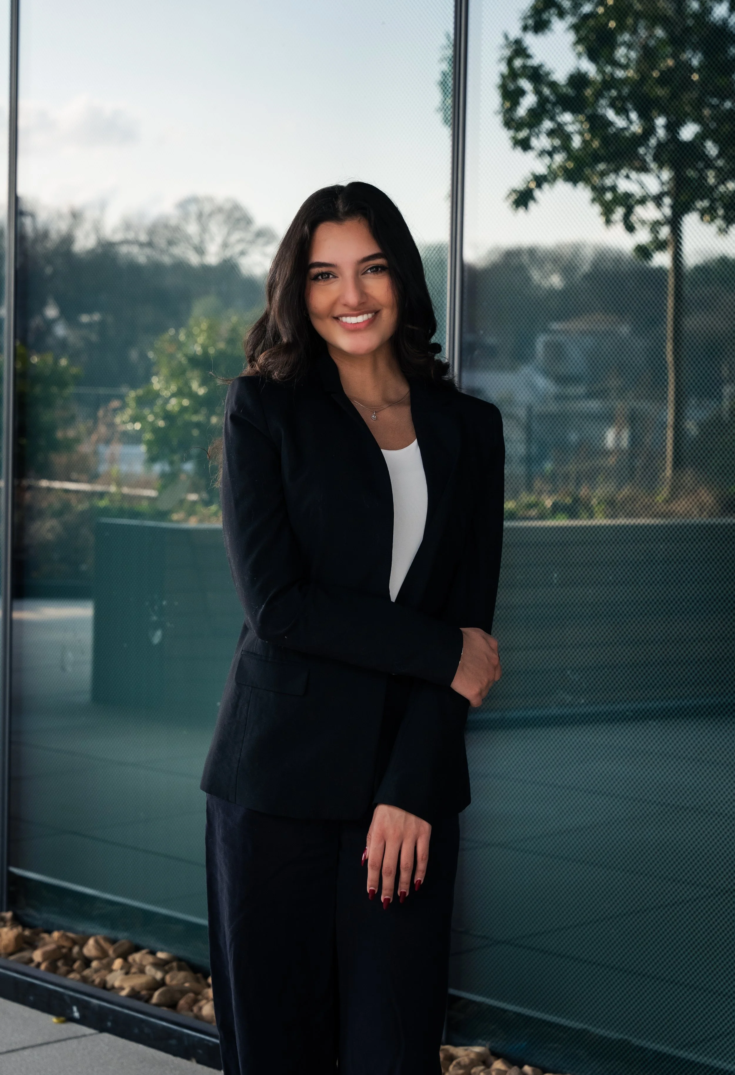 A woman with dark hair, dressed in a black suit and white shirt, smiling and standing outdoors near a glass building with trees in the background.