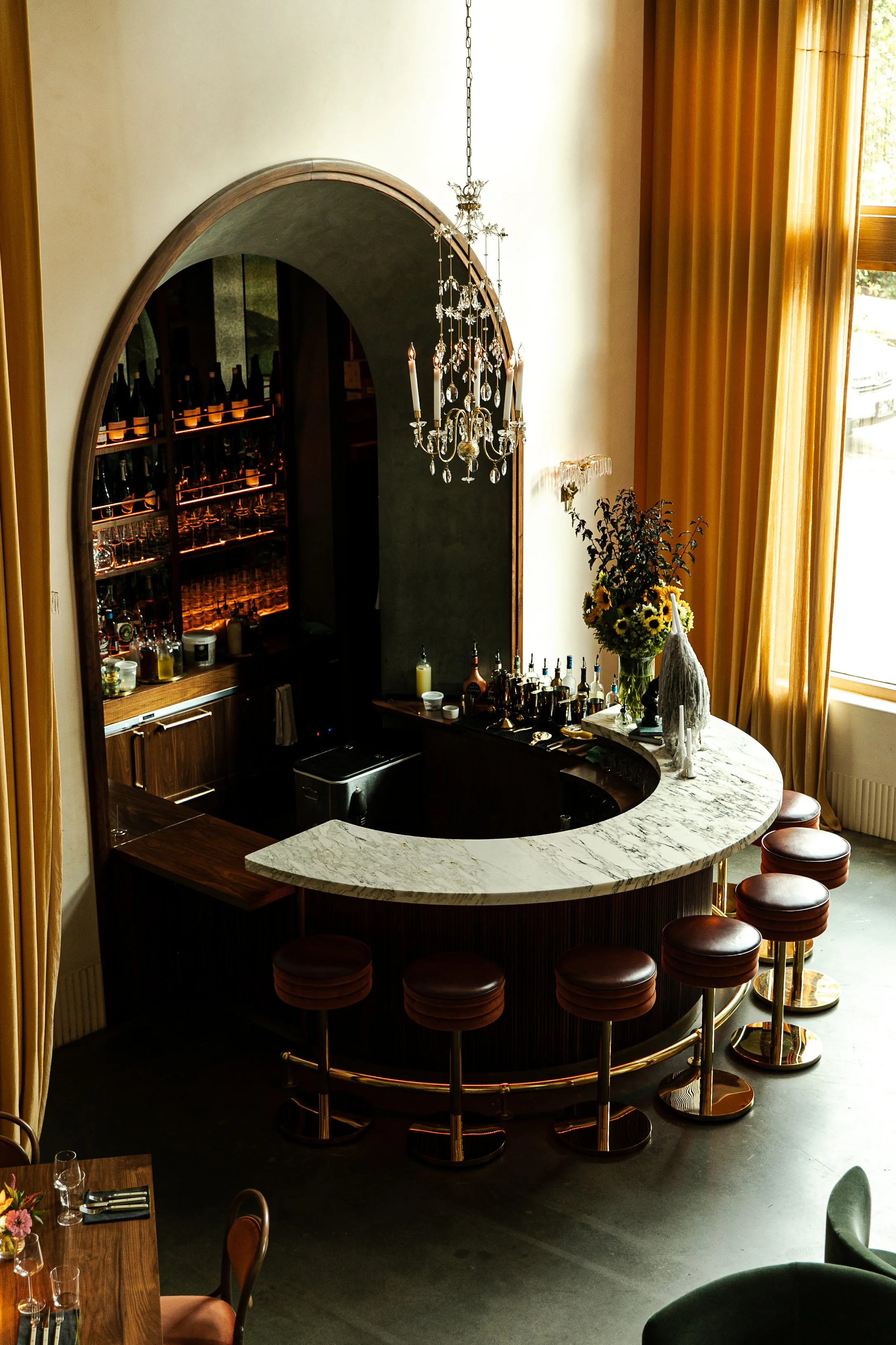 A restaurant interior with yellow curtains, a marble bar counter, bar stools, a chandelier, and a vase of sunflower flowers.