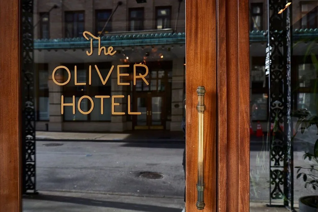 The glass door of The Oliver Hotel with its name written in gold lettering, wood framing, and a handle.