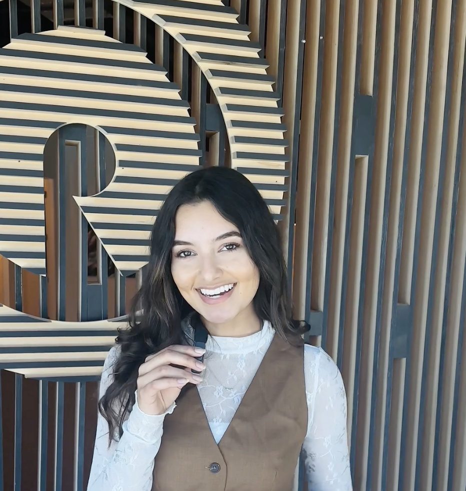 A woman with long dark hair smiling and holding a pen in front of a decorative wall with striped wooden slats.