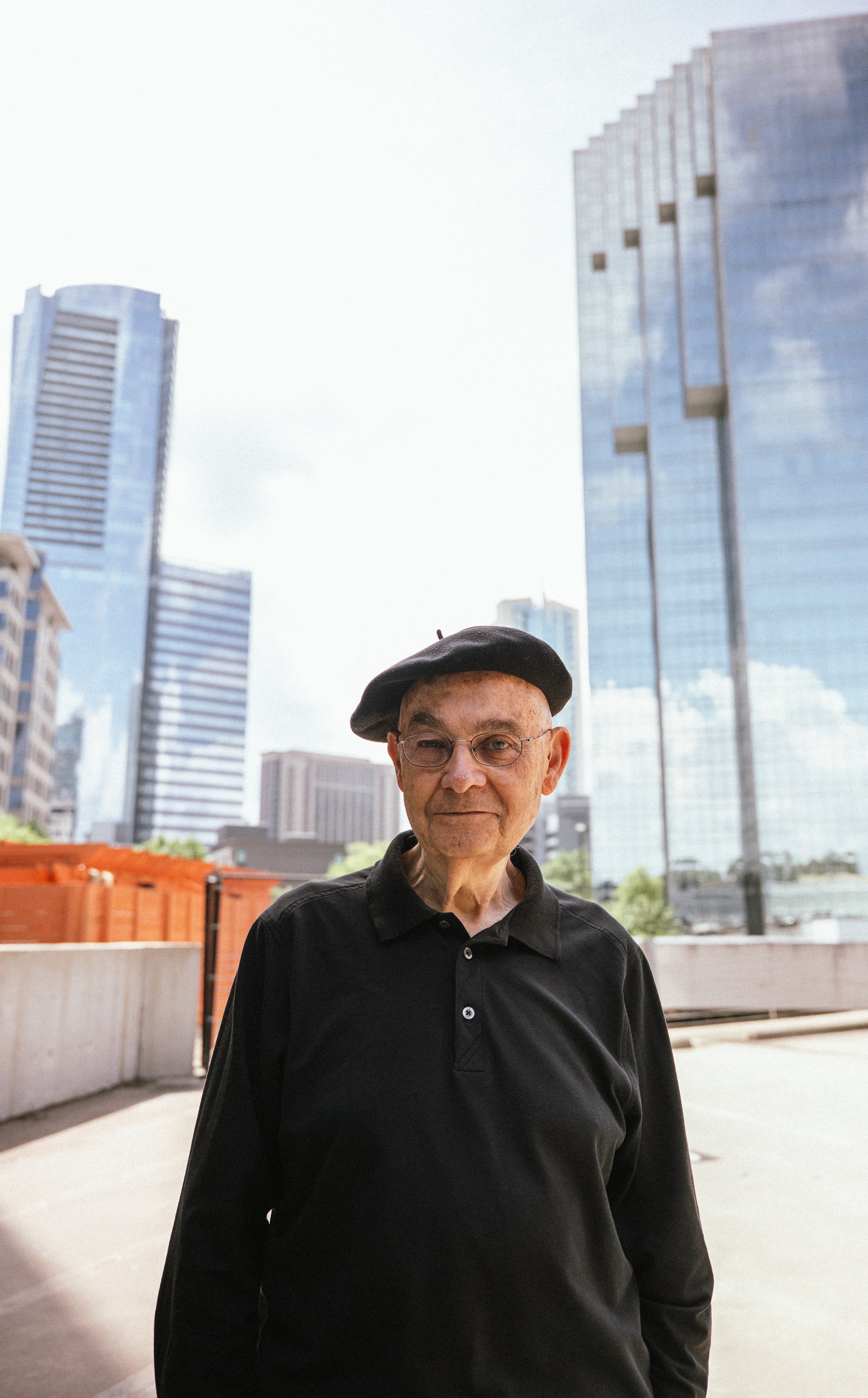 An elderly man wearing glasses, a black beret, and a black polo shirt standing outdoors in an urban setting with tall modern glass skyscrapers in the background.