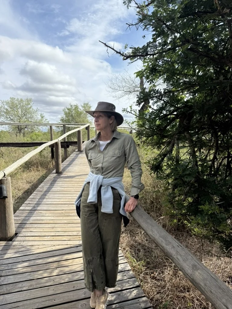 A woman in outdoor attire, including a wide-brimmed hat, standing on a wooden walkway next to a grassy area and trees.