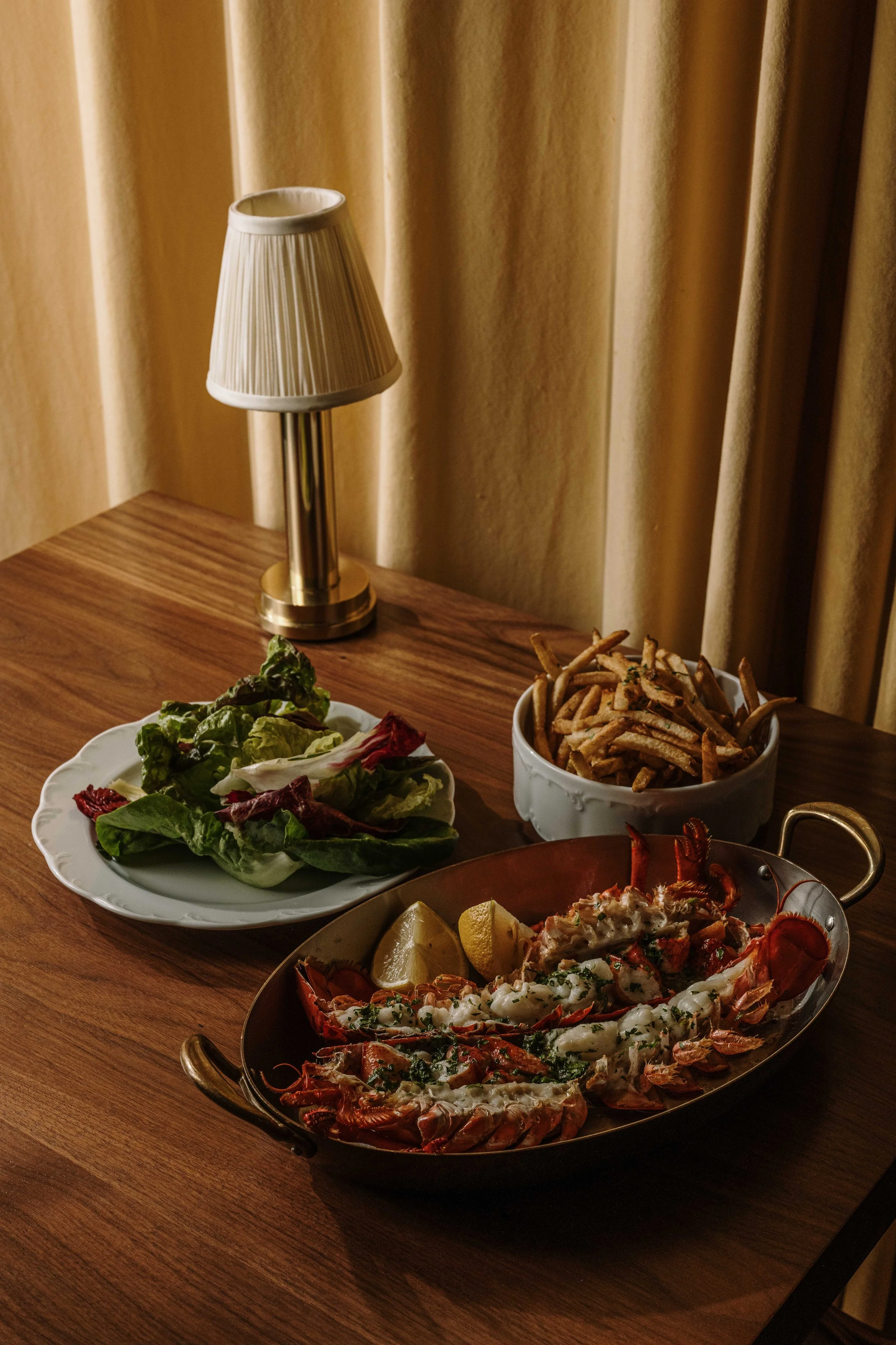 A seafood platter featuring lobsters, with lemon wedges, served on a rustic wooden table. In the background, a table lamp with a pleated shade and beige curtains are visible.
