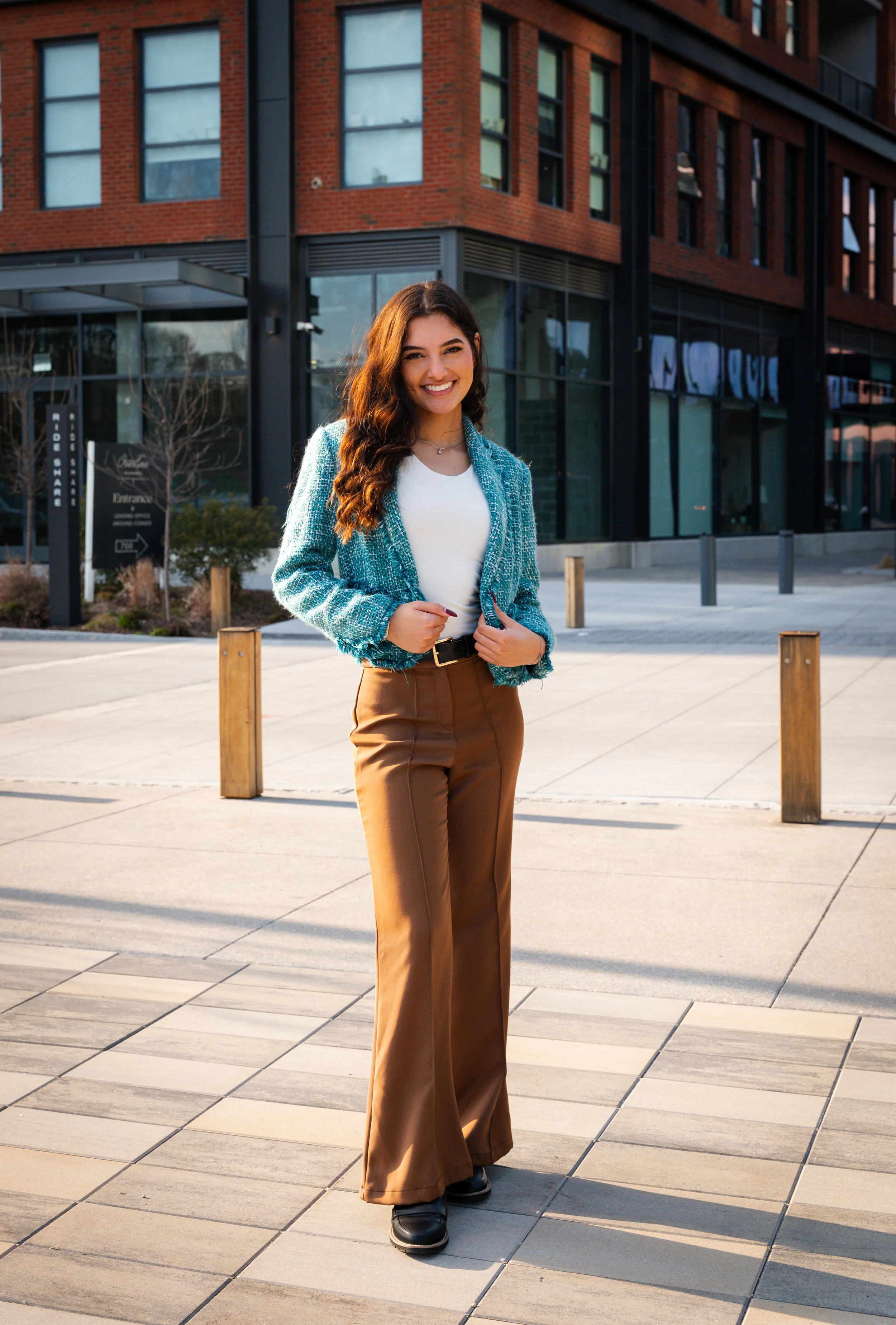 A woman with long brown wavy hair smiling and standing on a city sidewalk in front of a modern brick building with large glass windows. She is wearing a white top, teal blazer, brown high-waisted pants, and black shoes.