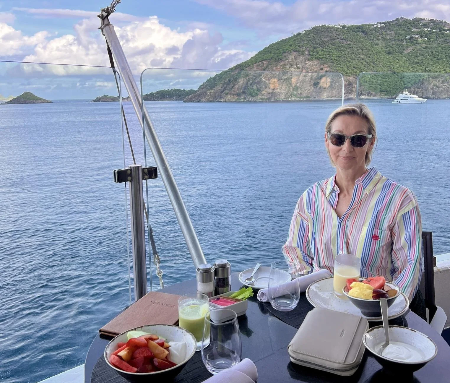 A woman in sunglasses and a striped shirt sitting at a table on a boat, with breakfast food including fruit and drinks, with a scenic ocean and hilly islands in the background.
