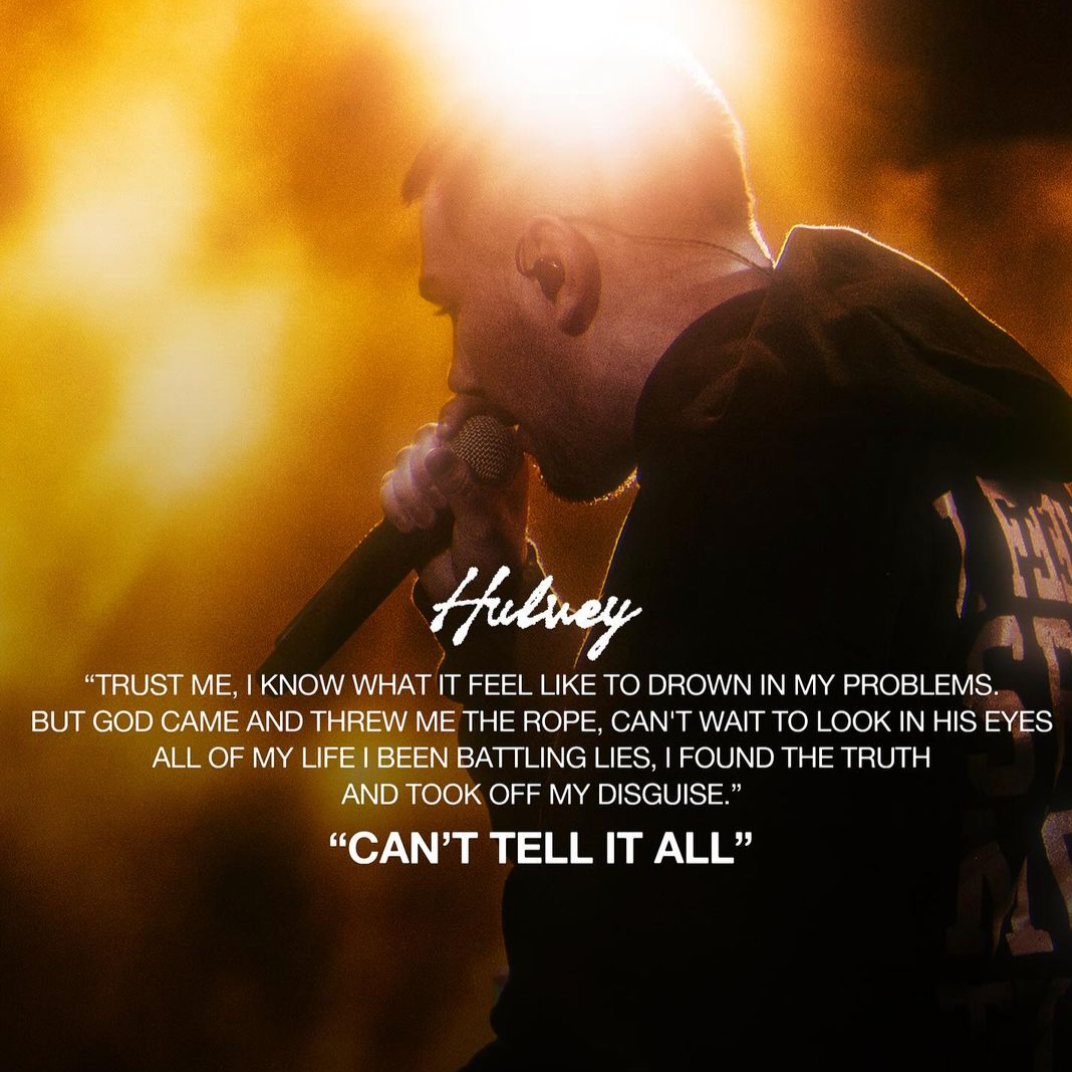 A man singing into a microphone on stage with intense lighting behind him, wearing a dark hoodie with white text.