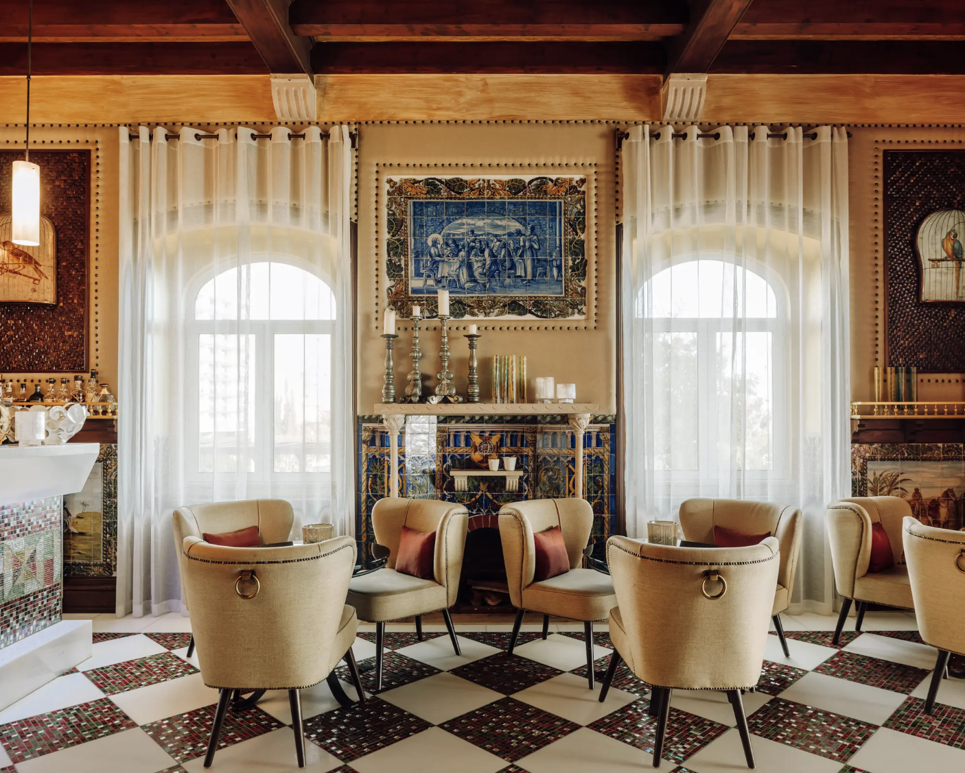 Elegant living room with beige armchairs, large arched windows with white curtains, a decorated fireplace, and framed artwork on the walls.