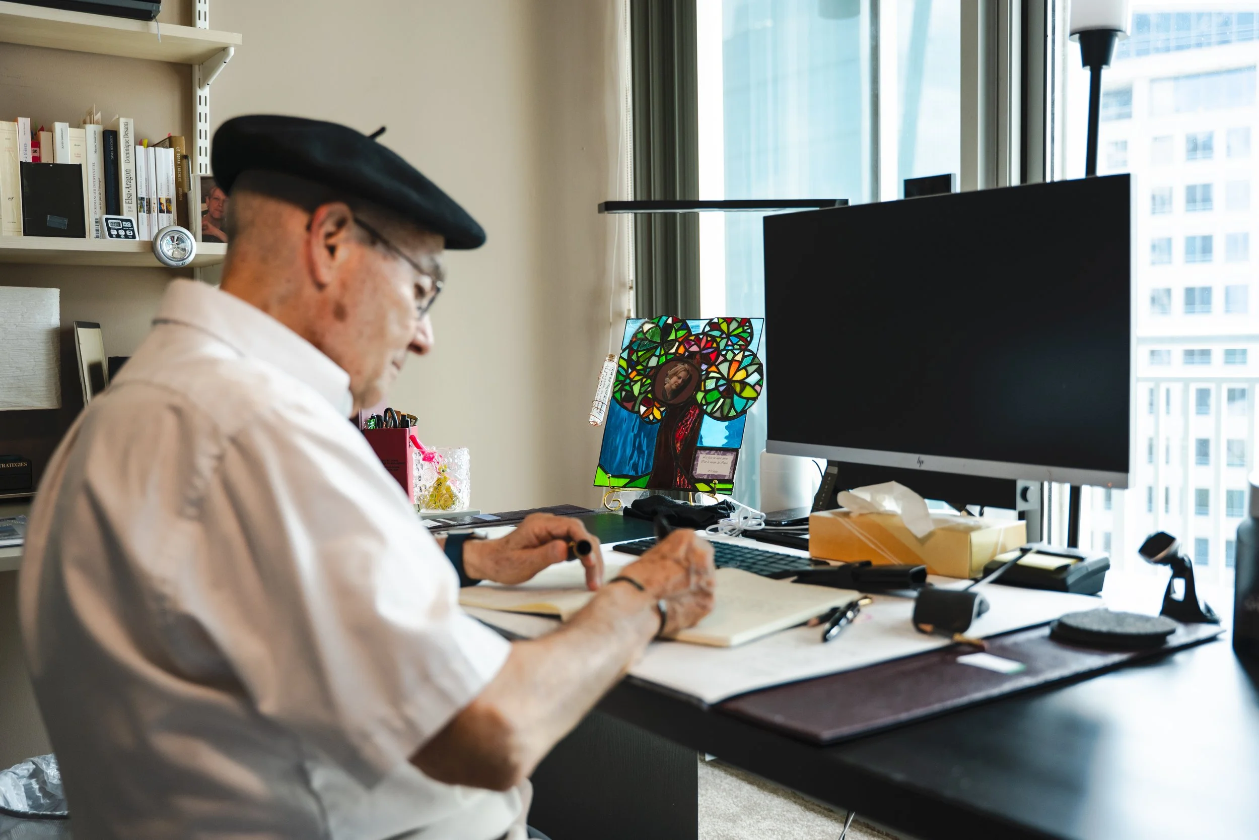 An elderly person sitting at a desk working on a notebook, with a computer monitor, colorful stained glass art, and writing supplies visible on the desk. There is a window in the background with a cityscape view.