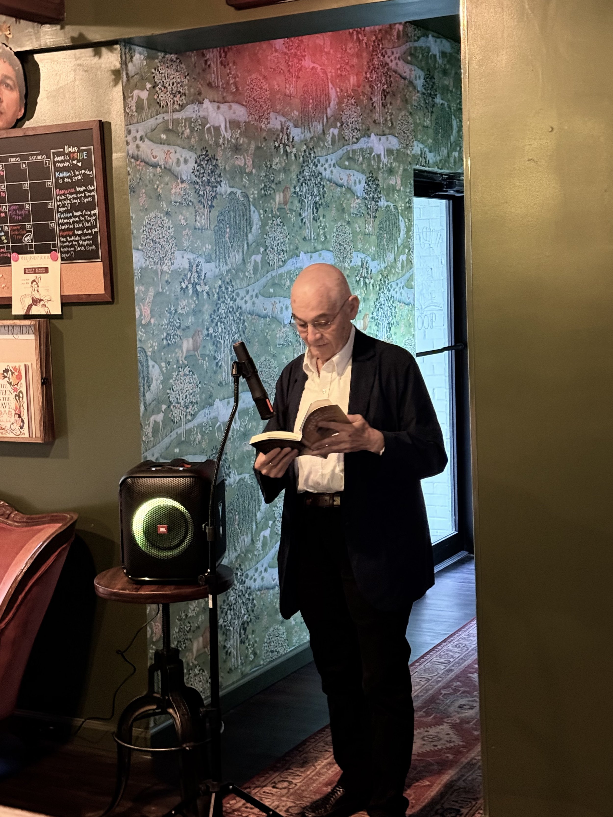 An elderly man with glasses, wearing a dark jacket and white shirt, is standing in front of a microphone, reading a book inside a room with a decorative mural of animals and trees on the wall behind him. There's a JBL speaker on a small wooden table near him.