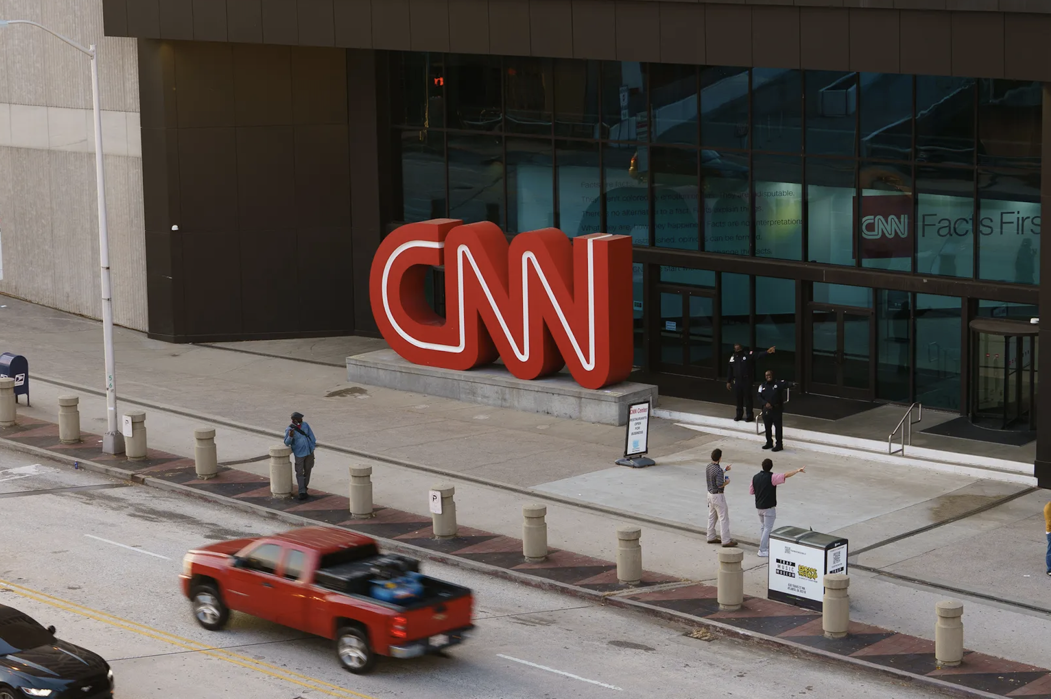 The exterior of a CNN building with a large red CNN logo outside. People are walking on the sidewalk, and cars are driving on the street in front of the building.