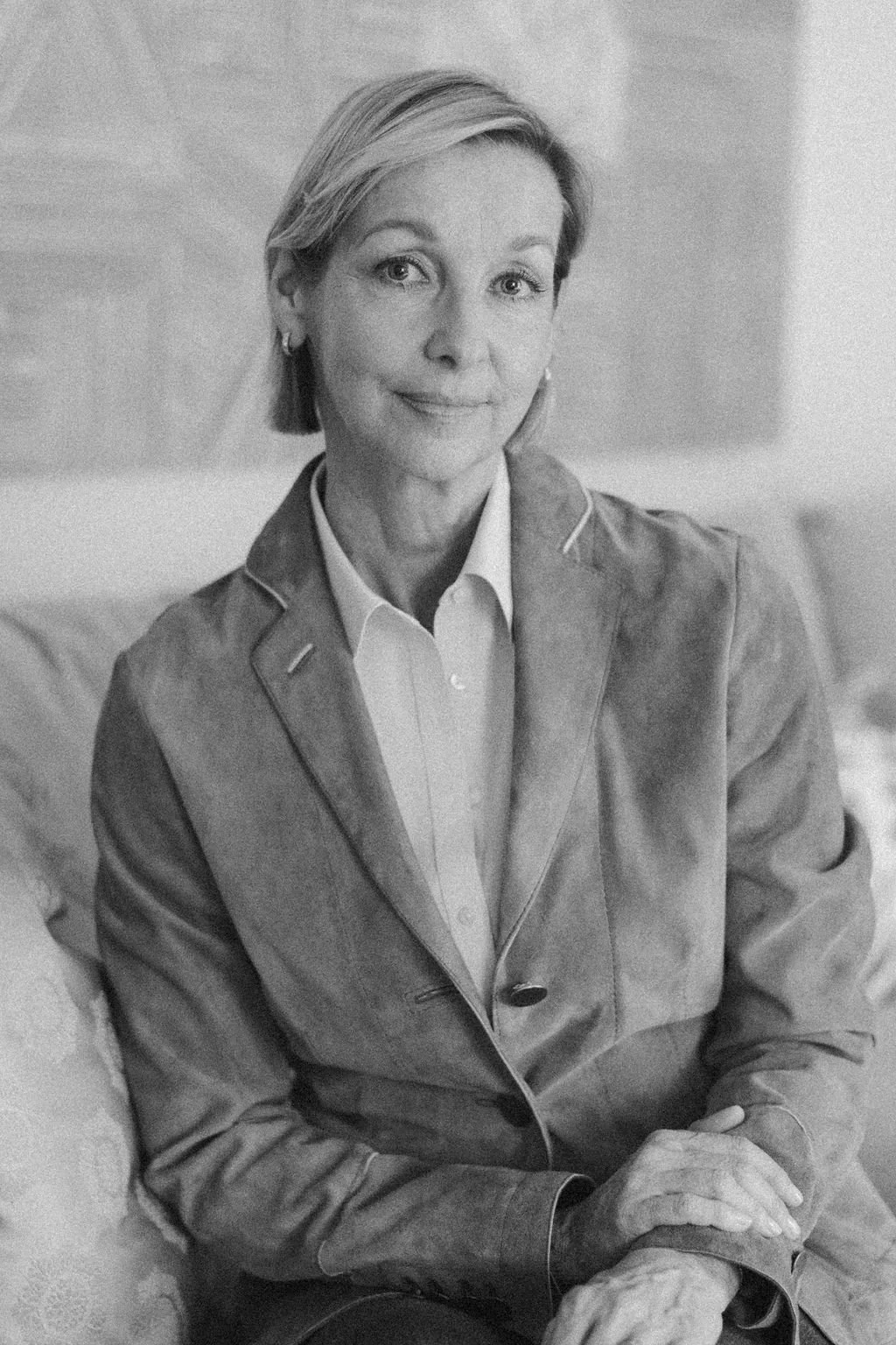 A black and white photo of a middle-aged woman with short hair, wearing a blazer and a collared shirt, sitting in a relaxed pose with her hand on her knee, smiling softly at the camera.
