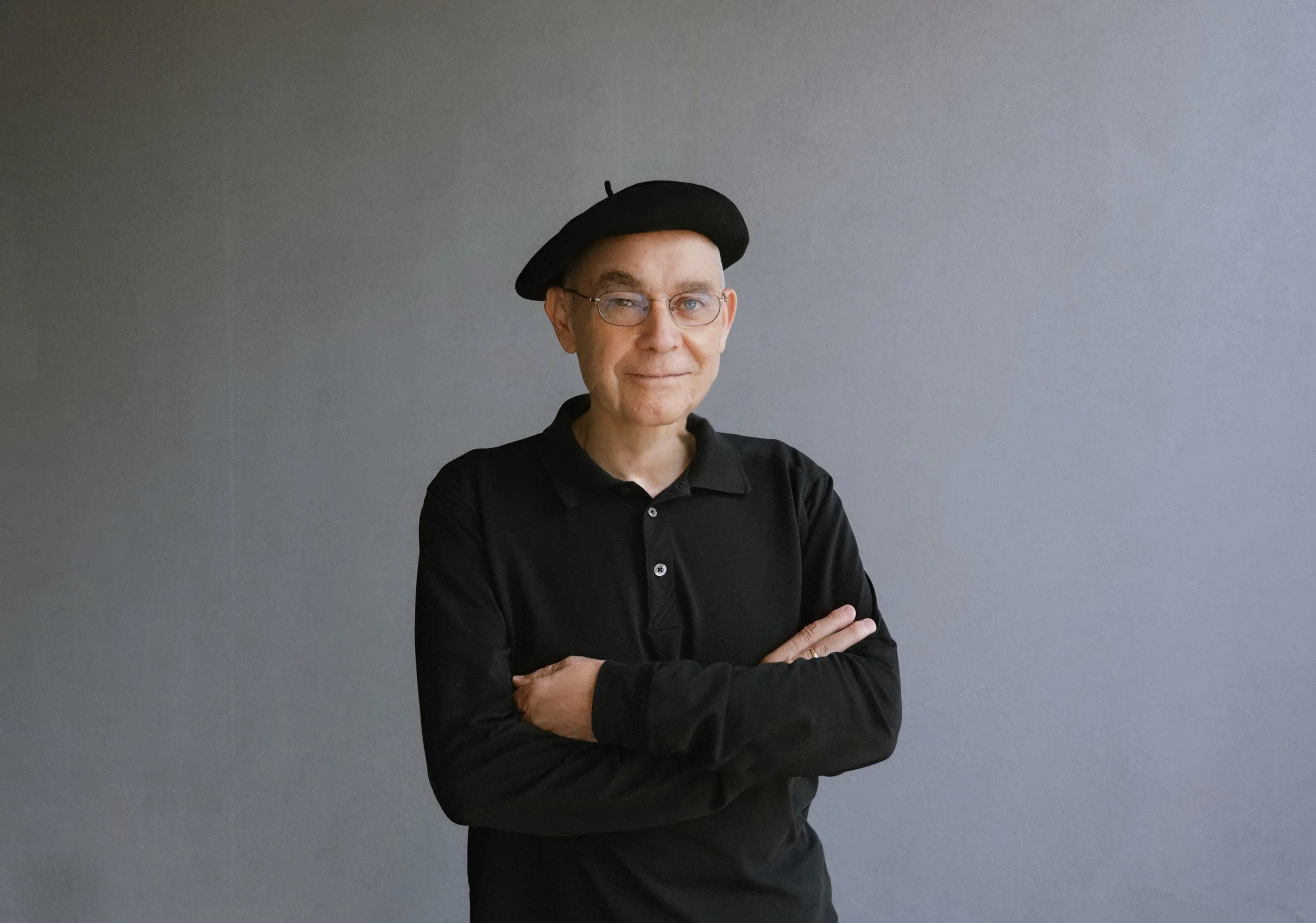 A man with glasses, wearing a black beret and a black shirt, standing against a plain gray background with arms crossed, looking at the camera.