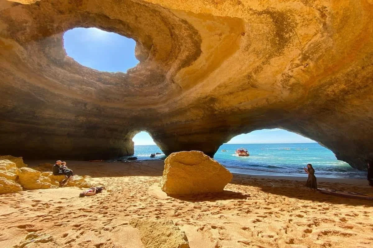 A natural sea cave with two large openings, showing the ocean and sky outside. The sandy beach inside the cave has rocks, and there are a few people relaxing and taking photos.