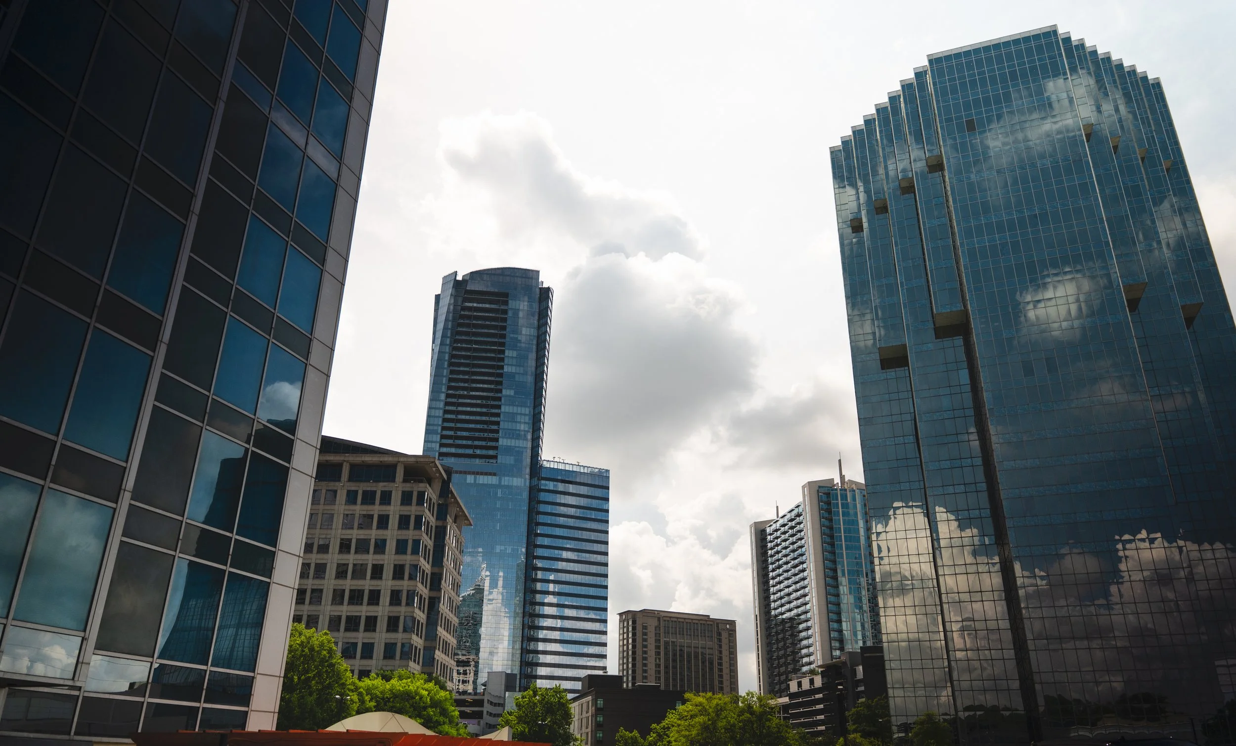 Modern glass skyscrapers reflecting the sky and clouds, with trees at the base.