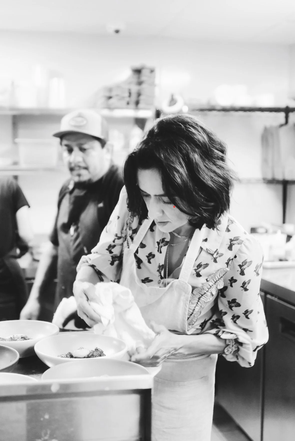 A woman in a butterfly print shirt and apron is preparing food in a kitchen, with bowls of ingredients in front of her, while a man in a cap looks on in the background.