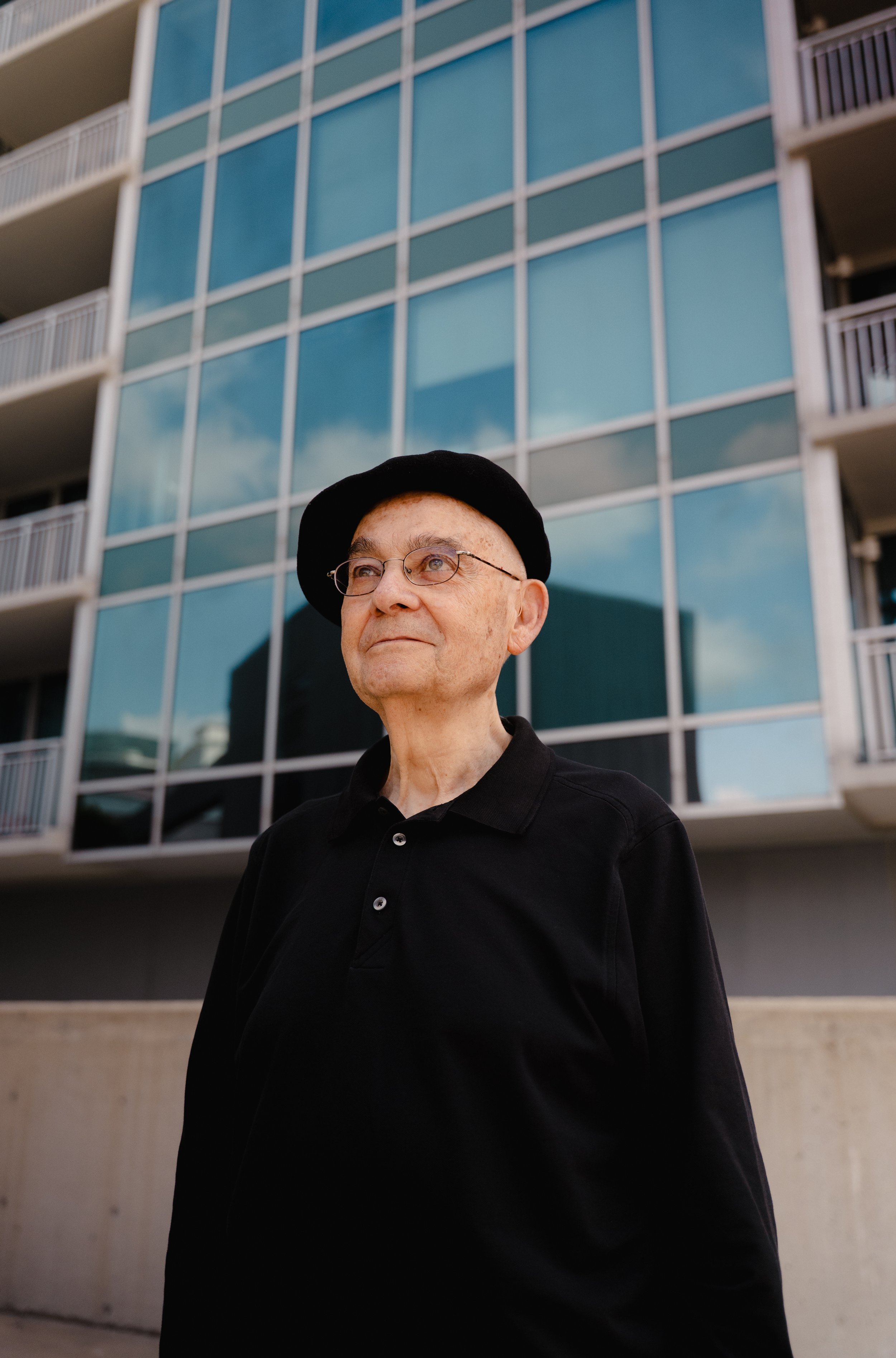 An older man wearing glasses, a black beret, and a black shirt stands outside in front of a modern glass building with balconies and reflections of the sky.