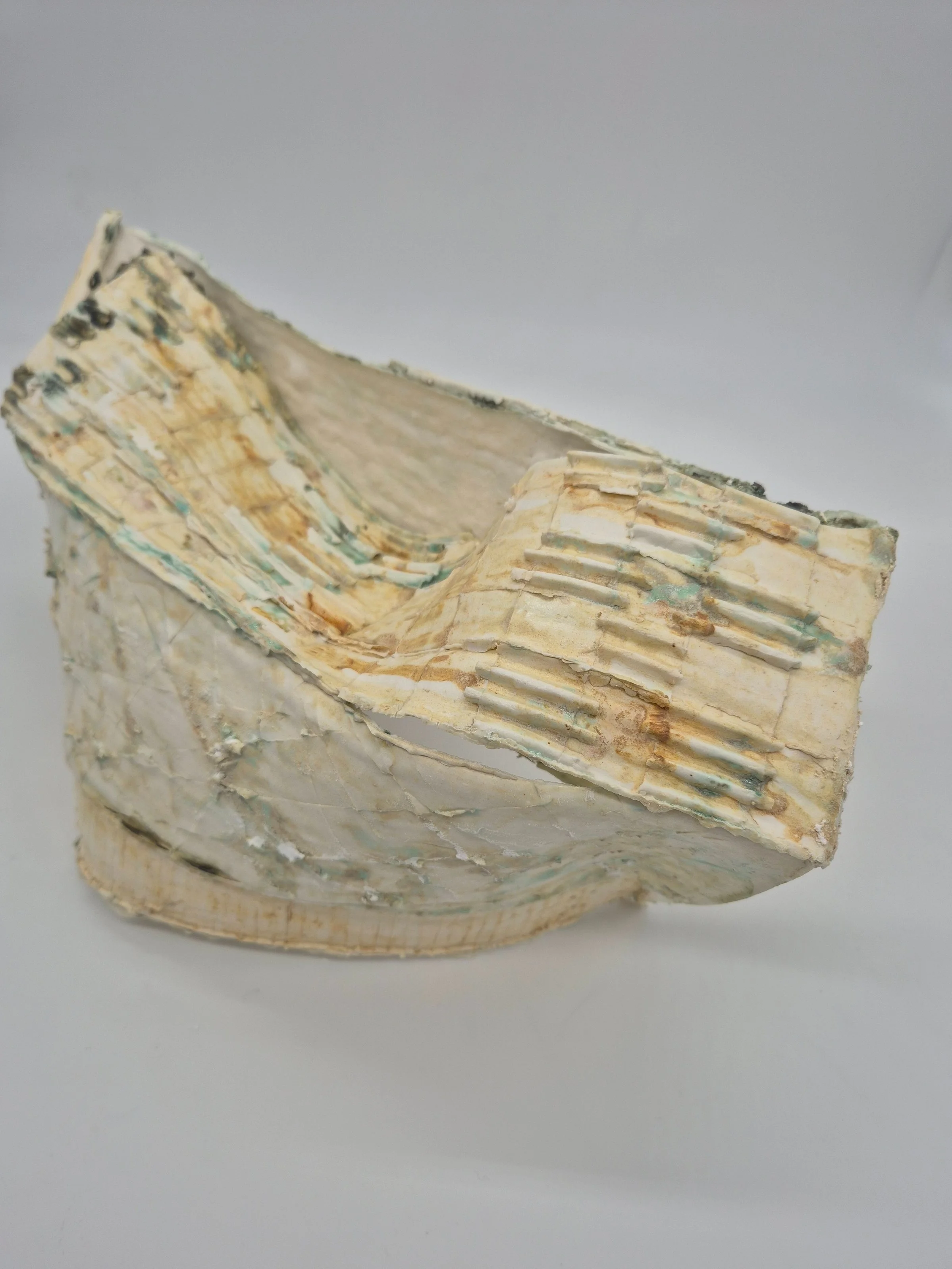 Abstracte keramische sculptuur met geometrische vormen en texturen in crèmekleur en groen-tinten, tegen een neutrale achtergrond.