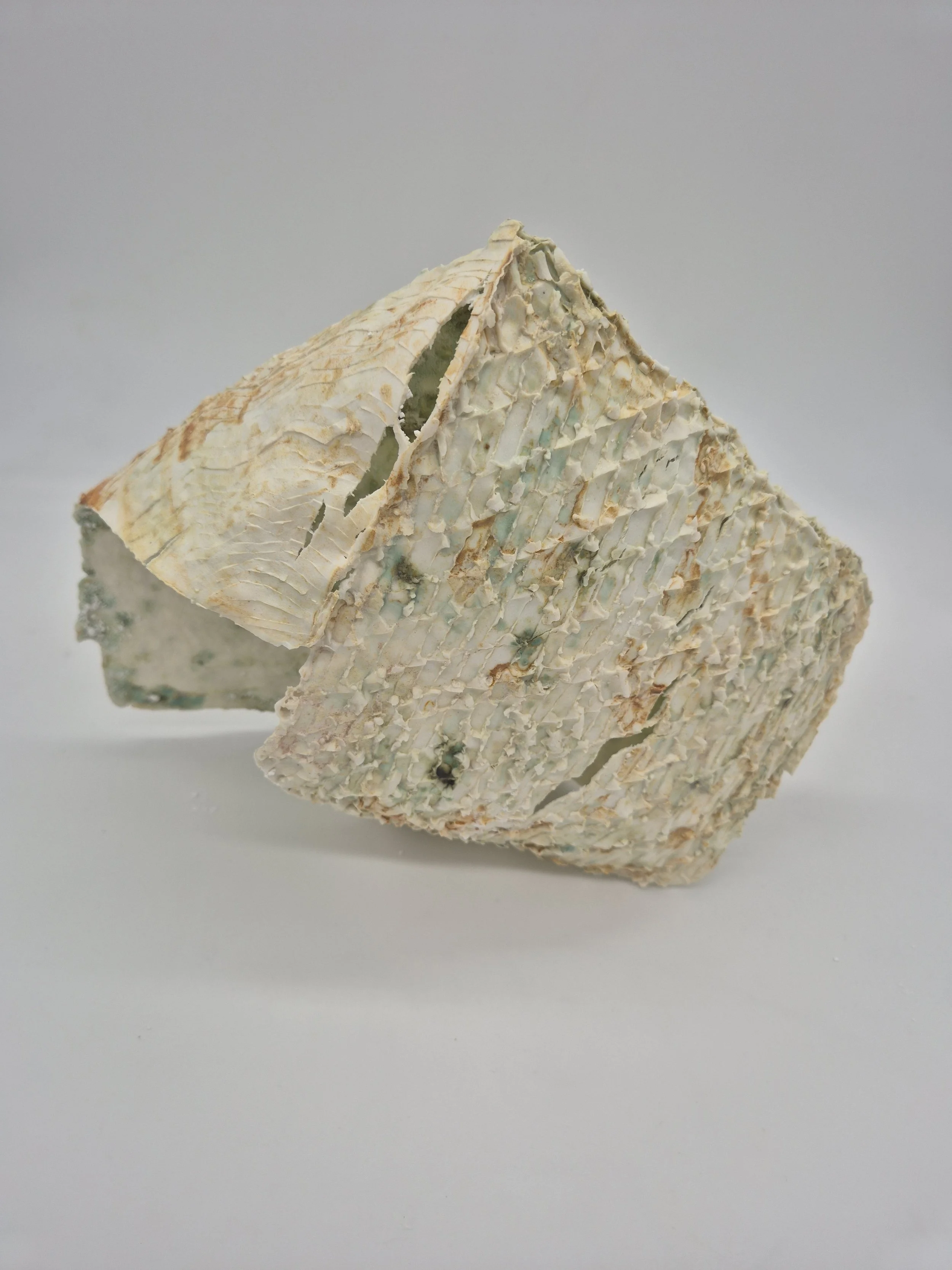 Een object met textuur, gemaakt van klei of keramiek, met een gebroken, rustieke uitstraling. De kleur is overwegend crème met lichte groene en bruine tinten.