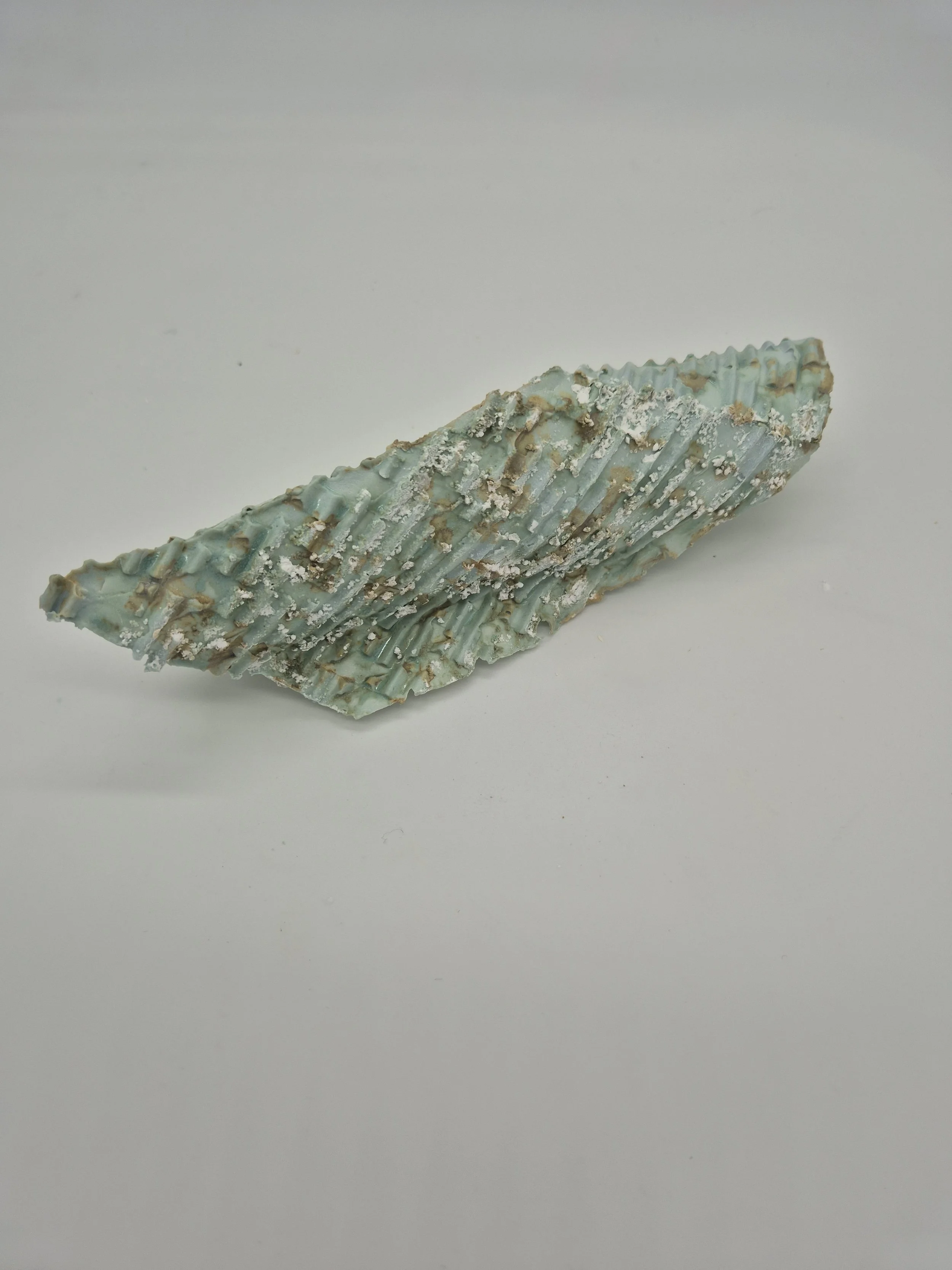 Een abstract kunstwerk met een geribbelde kopergroene textuur, bezaaid met witte en bruine accenten, op een witte achtergrond.