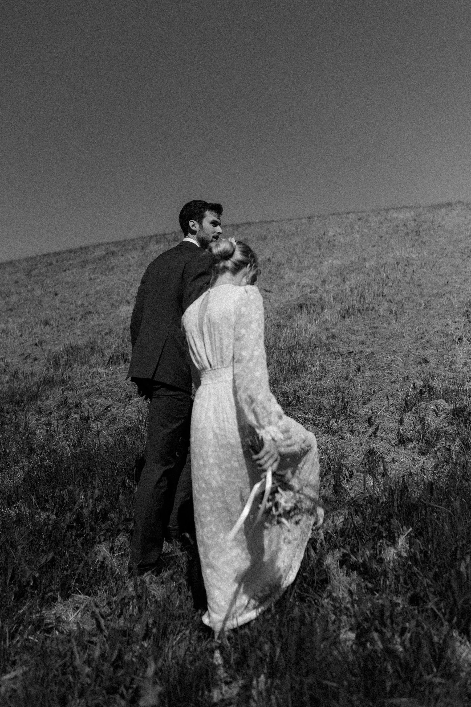 Black and white photo of a couple in formal attire walking up a grassy hill
