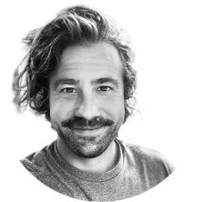 Black and white photo of a man with curly hair, a mustache, and a beard, smiling at the camera.