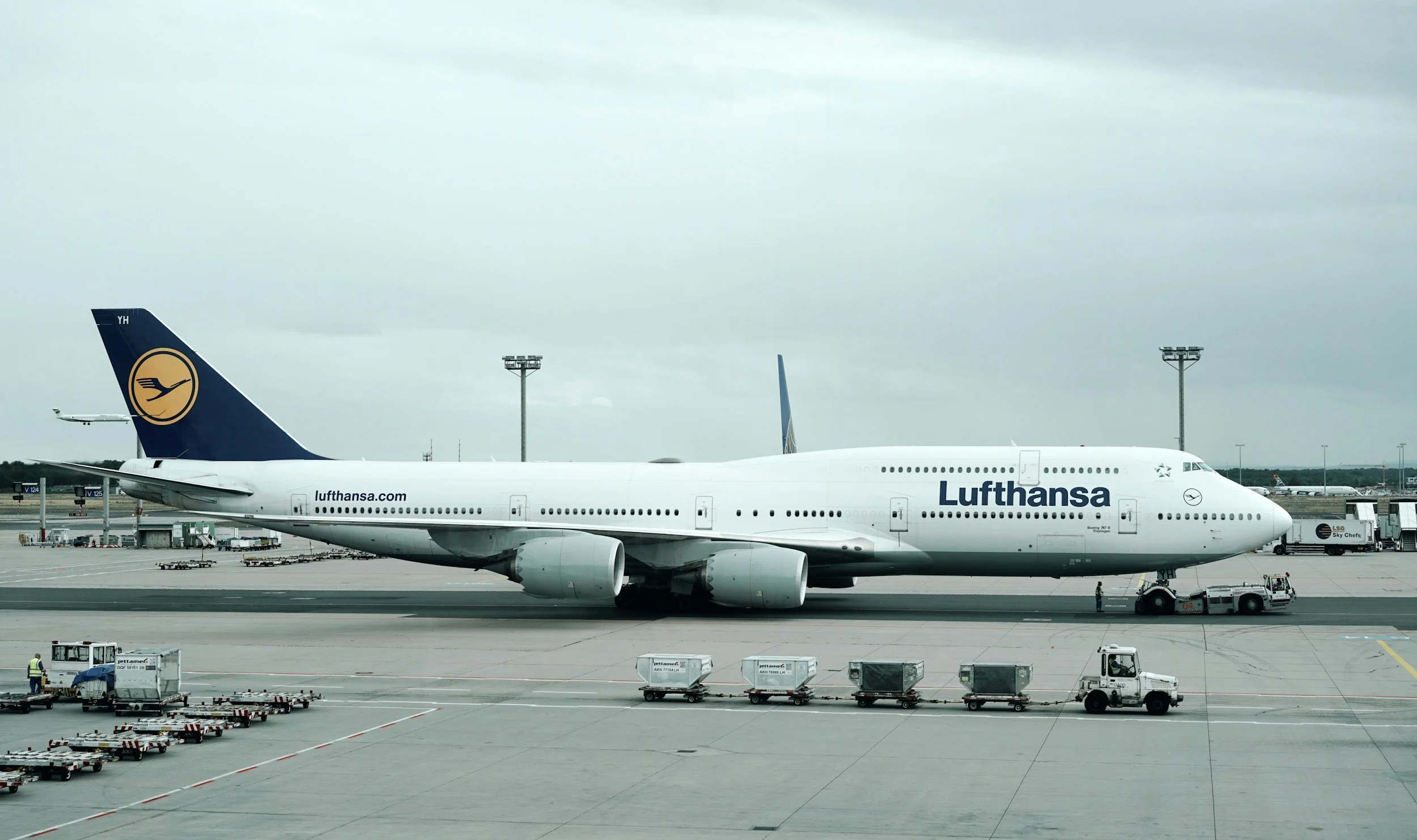 A Lufthansa Boeing 747 airplane parked at an airport gate, with luggage carts and ground crew nearby.