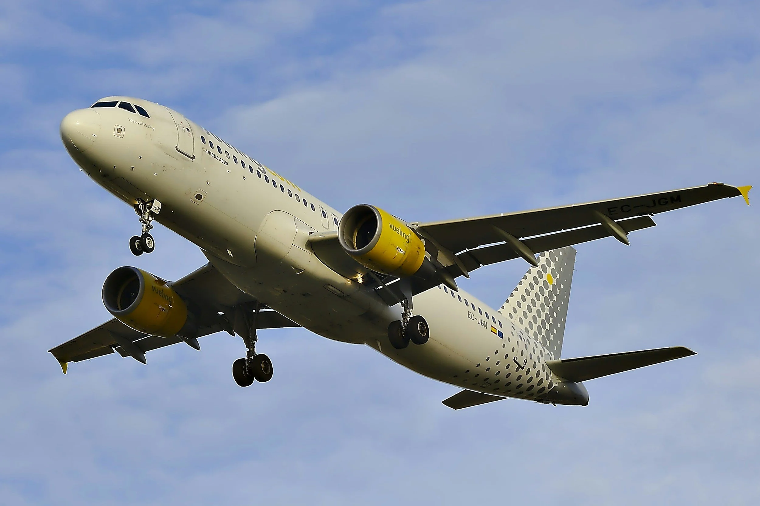 An Airbus A320 airplane in flight with landing gear down against a blue sky with scattered clouds.