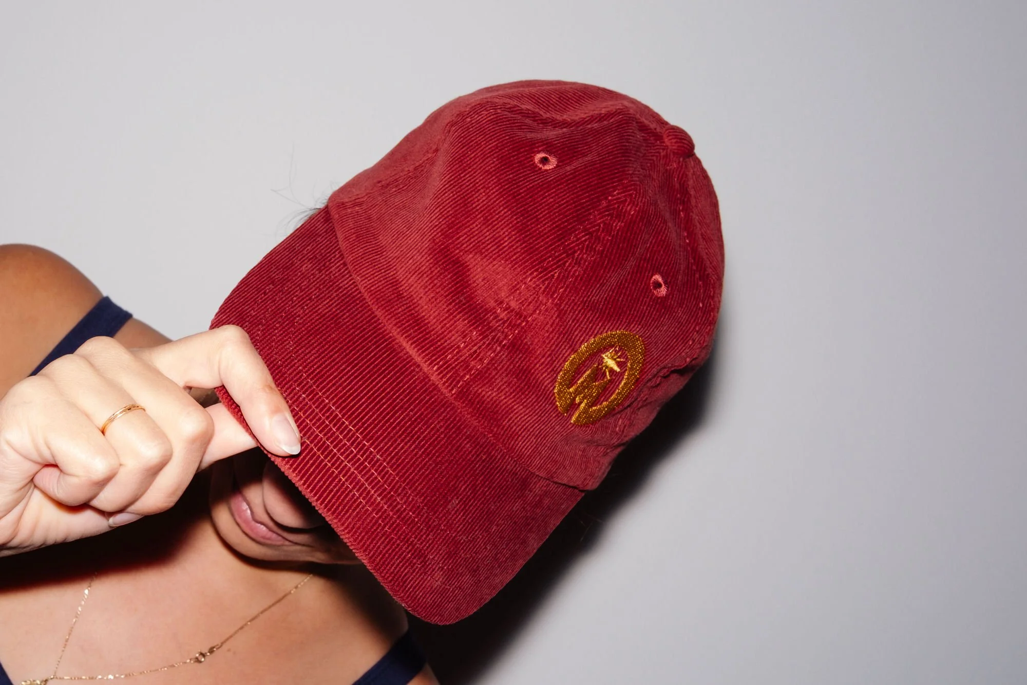 Person holding a red corduroy baseball cap with a yellow embroidered logo, partially covering their face, against a plain background.