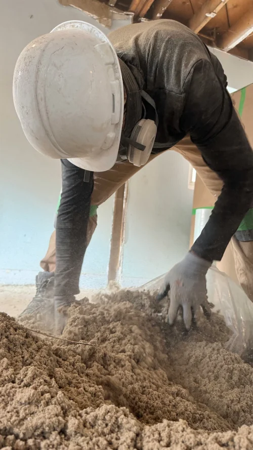 Worker in hard hat clearing loose insulation from a room under renovation