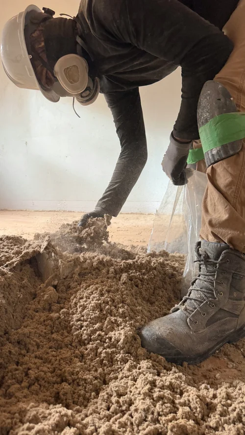Worker in hard hat and respirator shovelling old insulation by hand in a gutted room