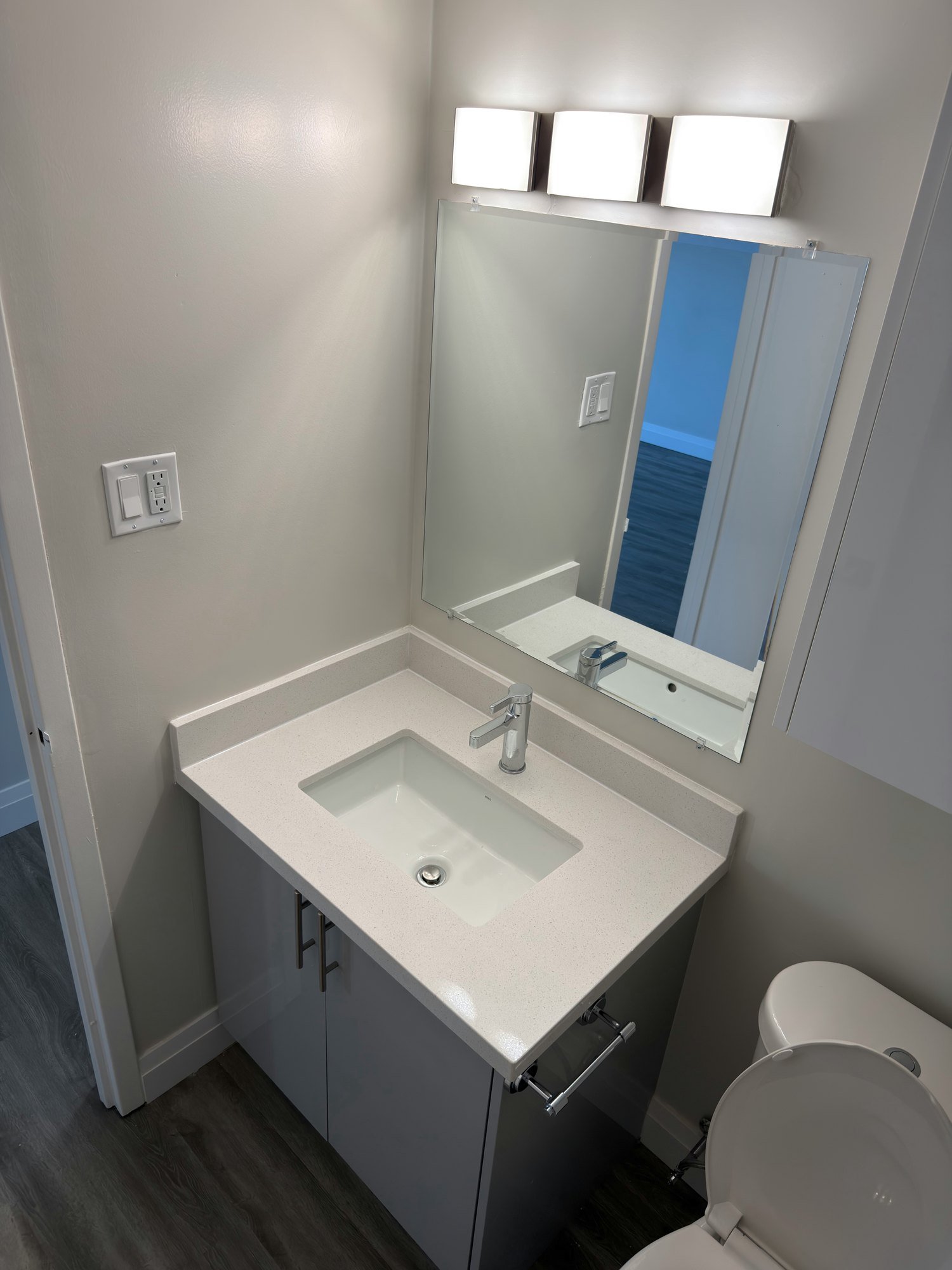 Finished bathroom corner with a grey vanity, a mirror with three wall lights, a wall cabinet and a white toilet