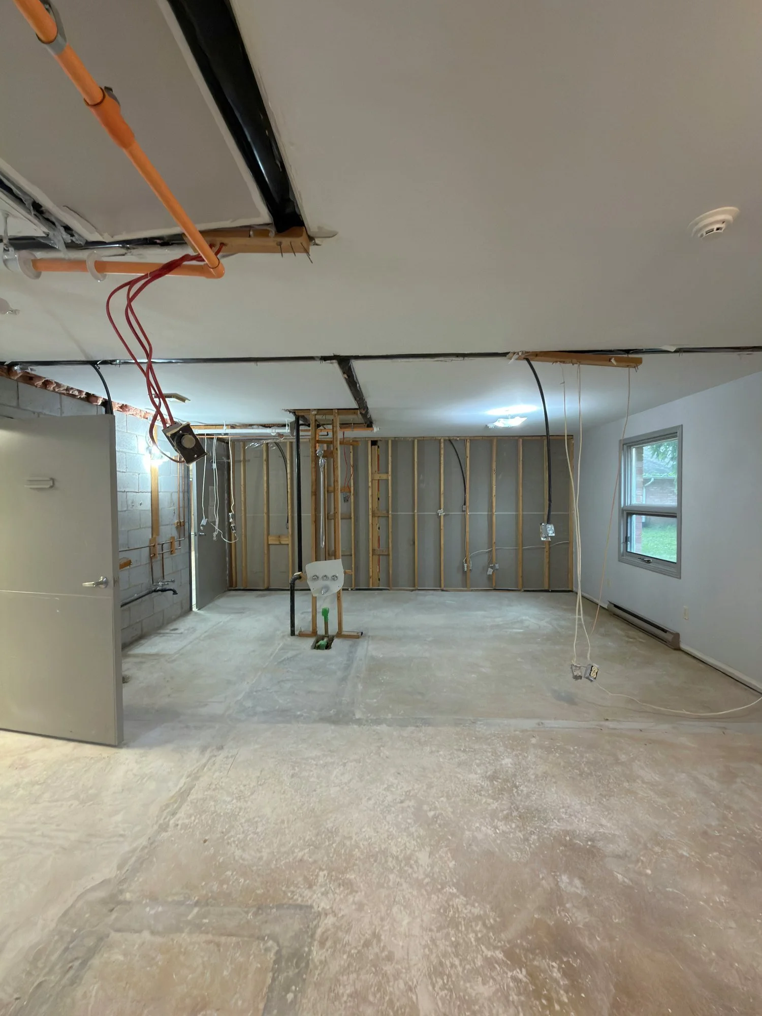 Room stripped to wood studs with pipes and wires exposed above and a bare floor
