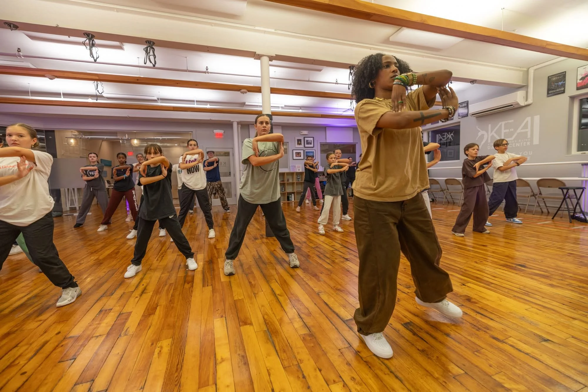 Dance class with diverse students exercising in a studio with wooden floors, led by an instructor.