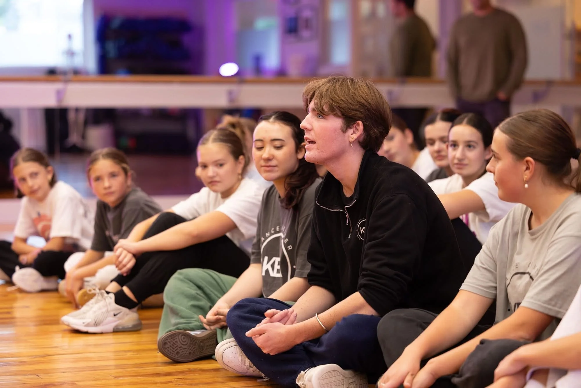 A group of children sitting on the floor in what appears to be a dance studio, with a young man speaking in front of them.