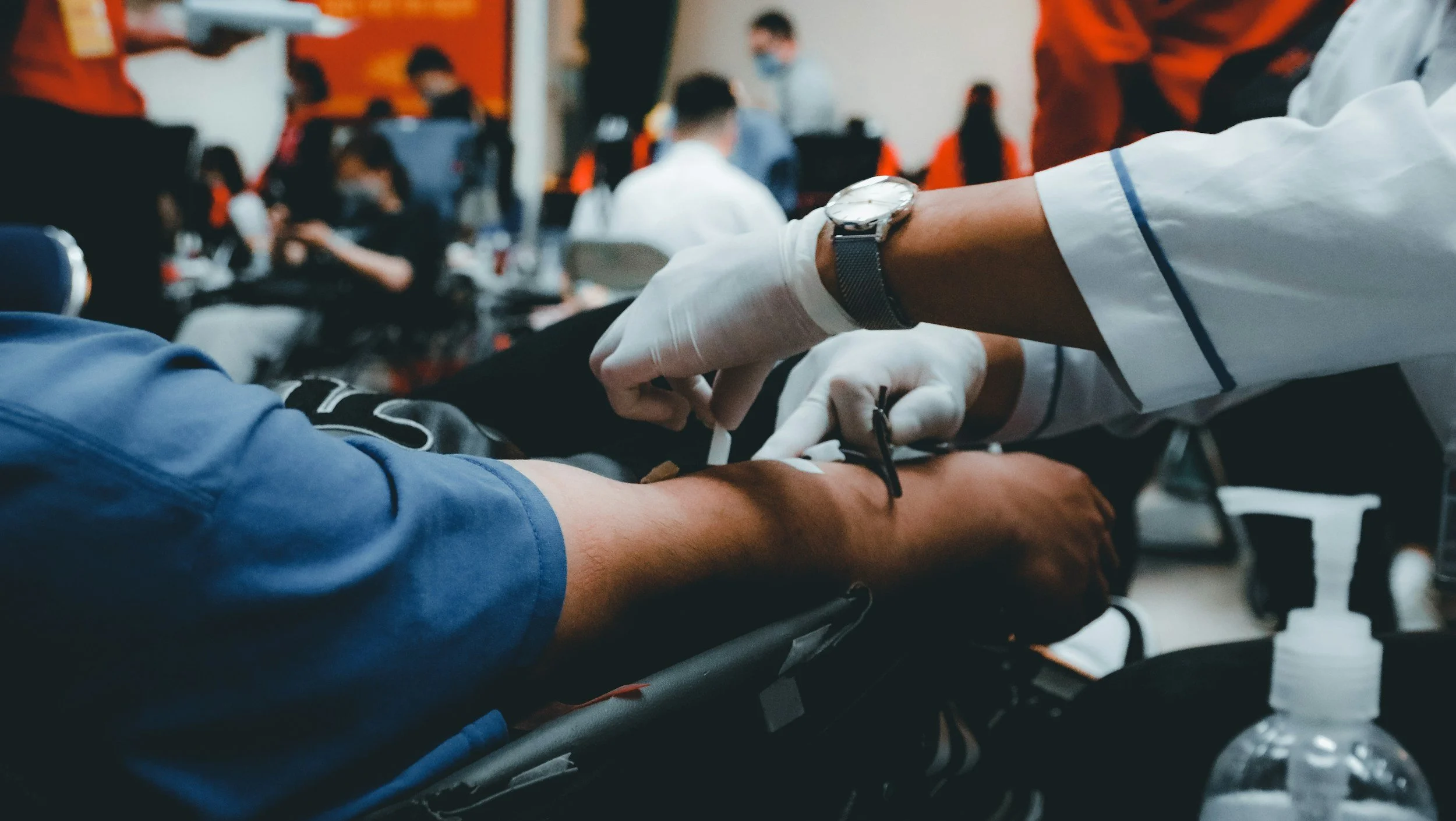 Healthcare workers in white gloves drawing blood from a patient's arm in a medical setting, with other seated patients and healthcare staff in the background.