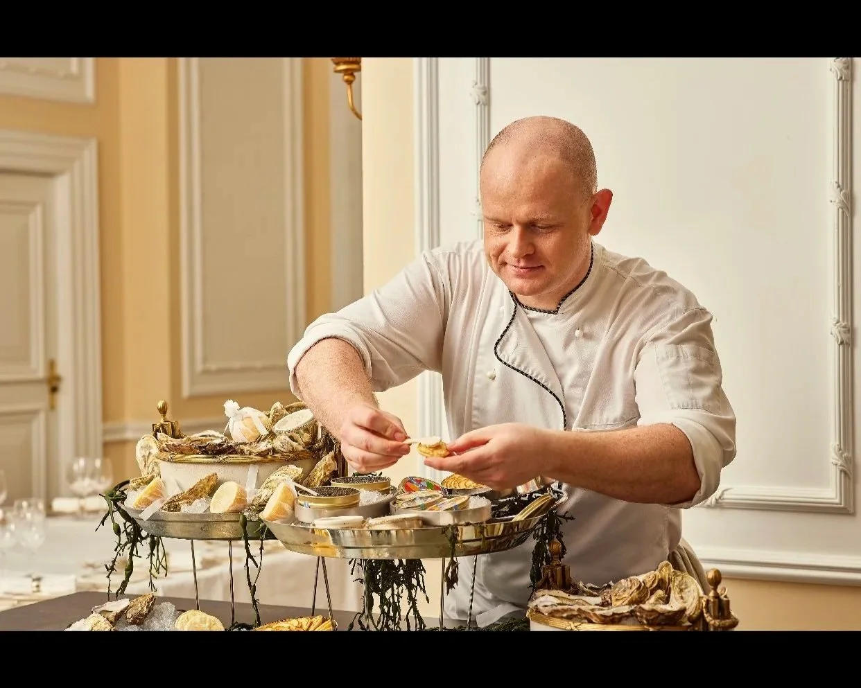 Chef with a bald head arranging oysters on a tiered tray in a decorated dining room.