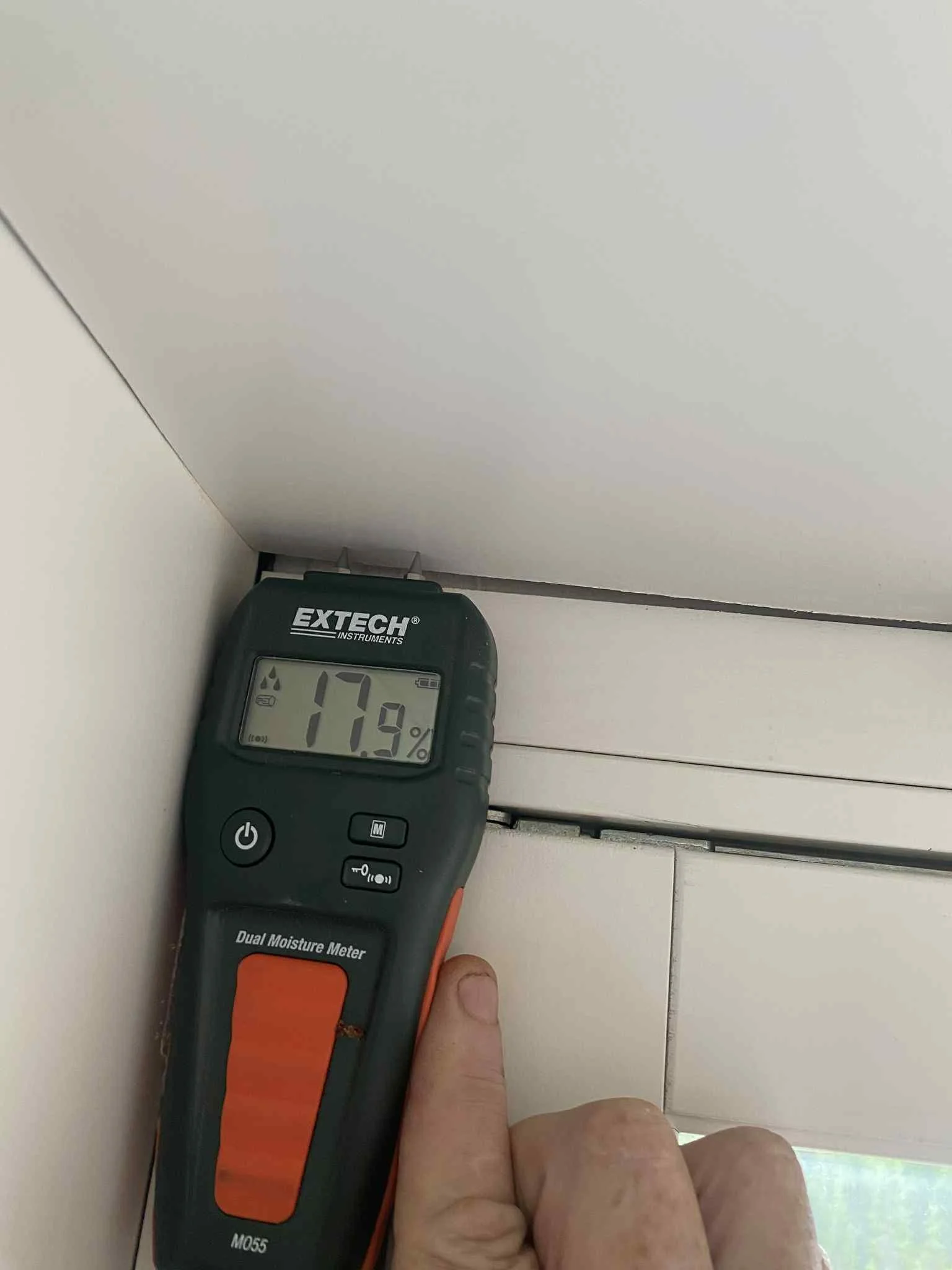 En person holder en kombinert fuktmåler som måler 17,9 % i takhjørnet ved veggen.
