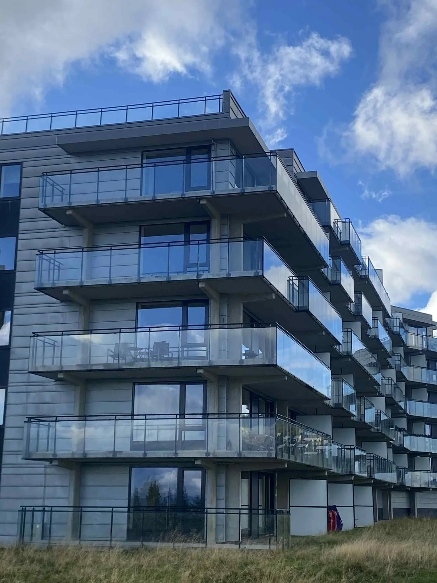 Et moderne leilighetsbygg i flere etasjer med glassbalkonger som speiler den blå himmelen og skyene. Bygningen er omgitt av et gressområde.