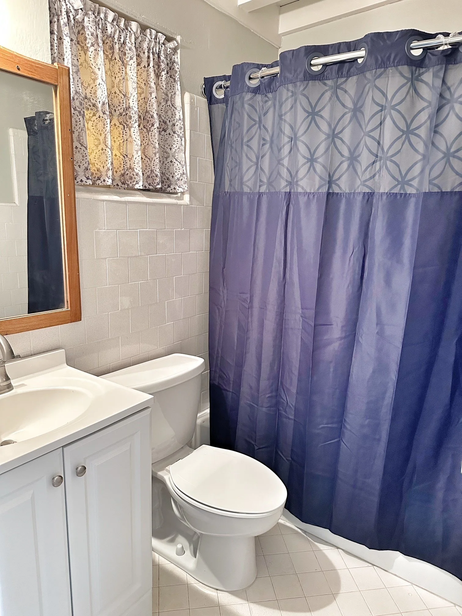 Bathroom with white vanity, toilet, mirror, window with patterned curtains, and a blue shower curtain.