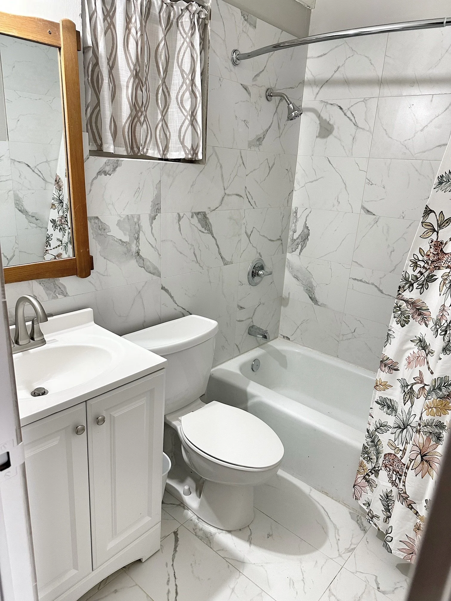 Small bathroom with marble tile walls and floor, a white toilet, a white vanity with a sink, a mirror with a wooden frame, a shower with a curved shower rod and patterned shower curtain.